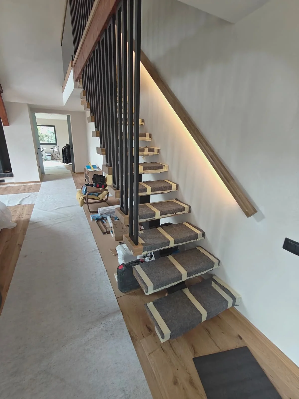 Wooden staircase with black metal rails under construction in a modern home, with tools and materials on the floor and a hallway leading to another room.