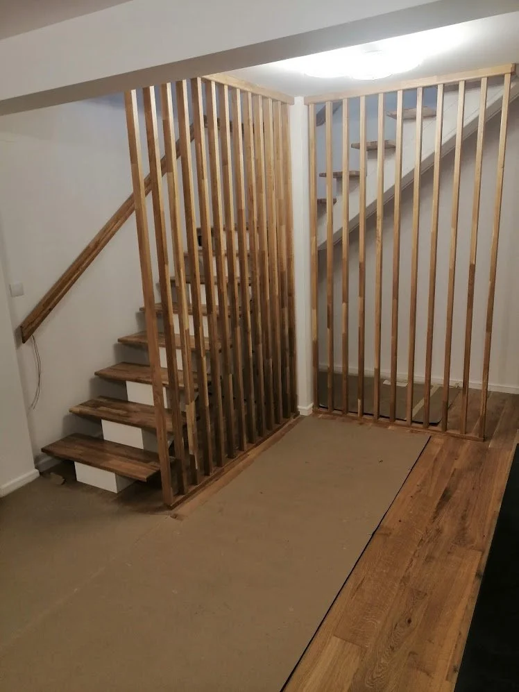 Wooden staircase with vertical slat railing under construction in a home, with some carpet and hardwood flooring.