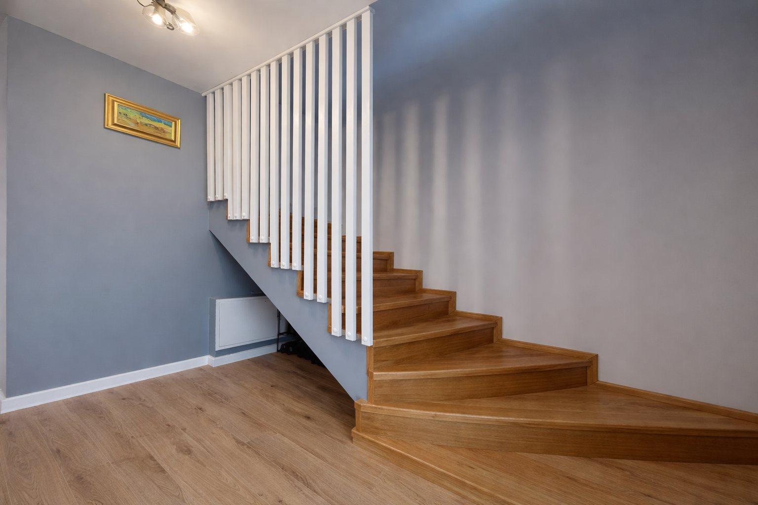 Interior view of a staircase with wooden steps and white vertical balusters, a gray wall with a framed landscape painting, and wooden flooring.