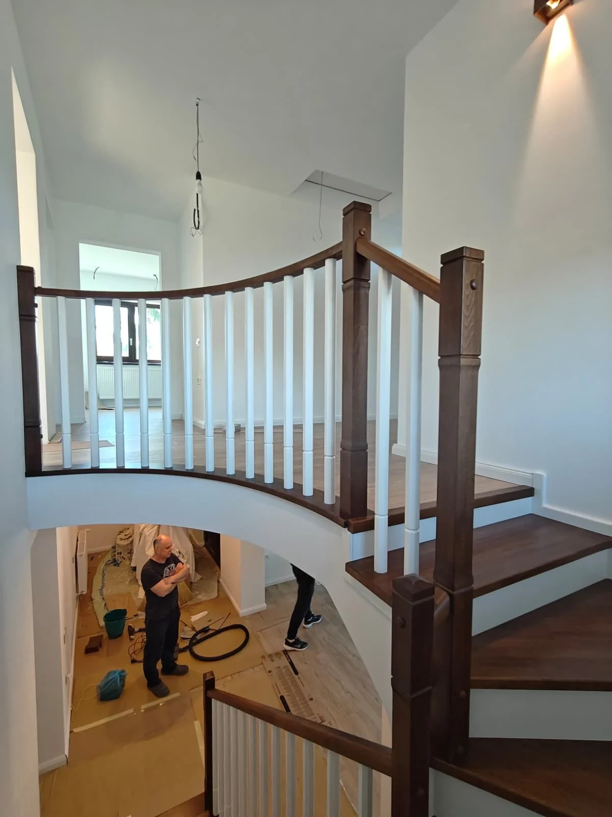 Interior of a home with a curved staircase featuring wood and white railing, with workers installing or finishing the space, including visible tools and materials on the floor.