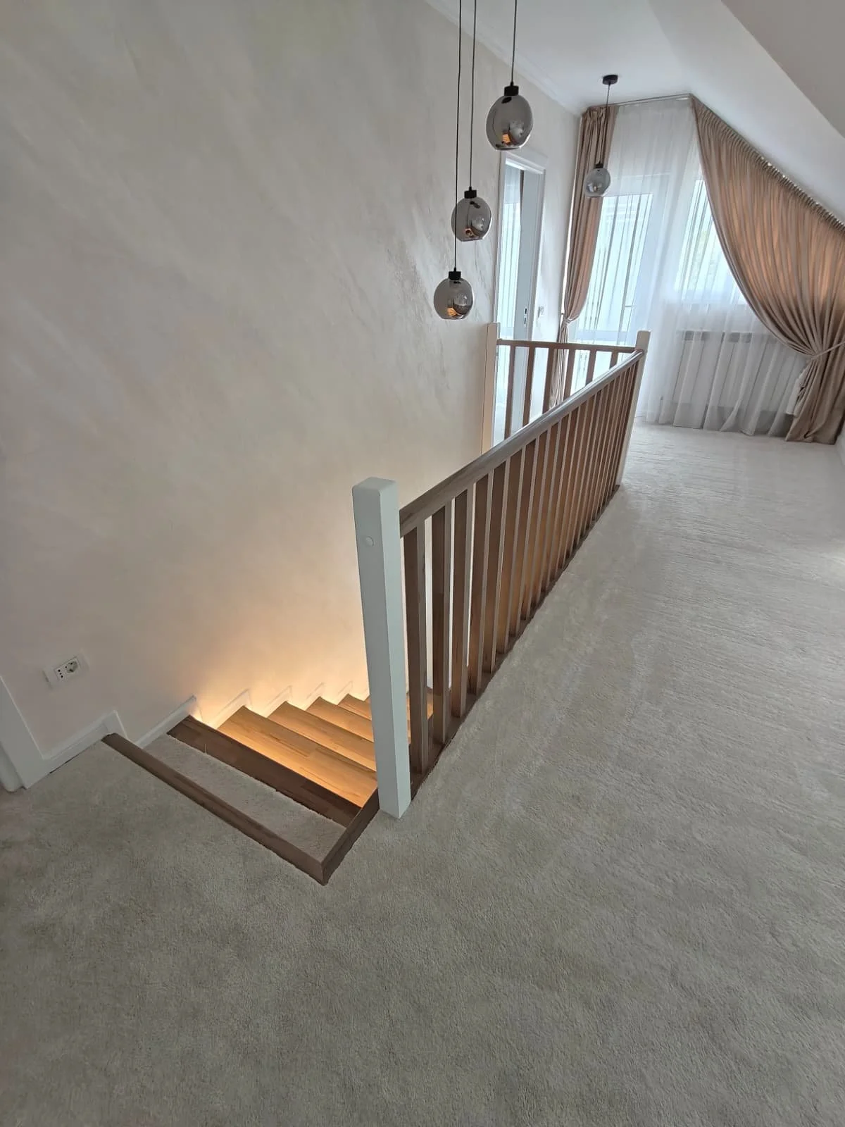 Interior view of a carpeted staircase area with a wooden railing, hanging pendant lights, large window with sheer curtains, and natural light.
