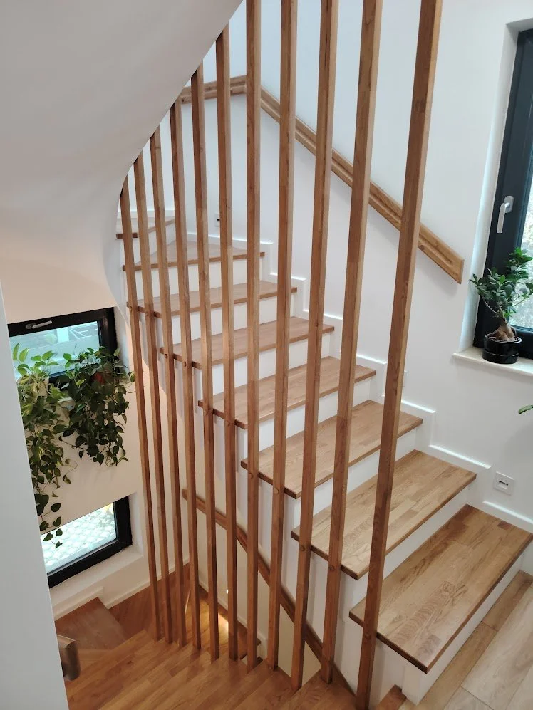 Interior view of a staircase with wooden steps, partially enclosed by vertical wooden slats, with white walls and a window with potted plants.