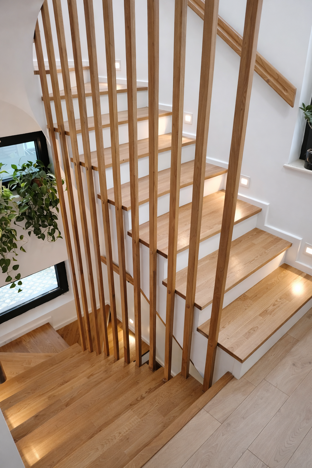 View of a staircase with wooden steps, a wooden railing, and modern wall-mounted lighting, next to a window with a potted plant on the windowsill.