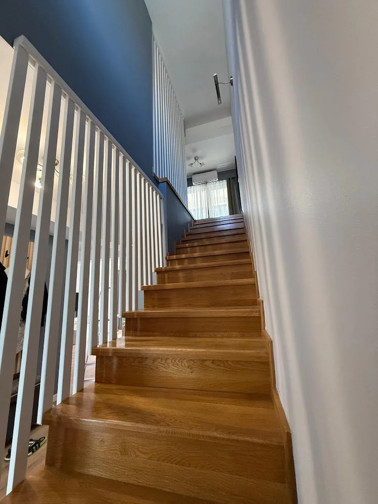 Wooden staircase with white railings on the left and white wall on the right, leading to a brightly lit area with curtains at the top.
