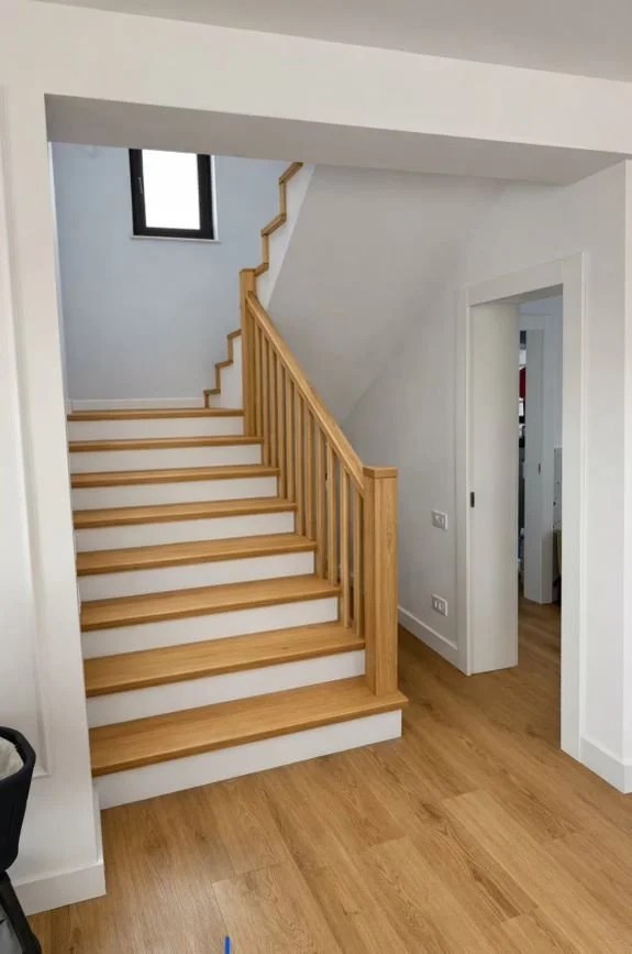 Wooden staircase with banister inside a modern home, adjacent to a room with white walls and a hardwood floor.
