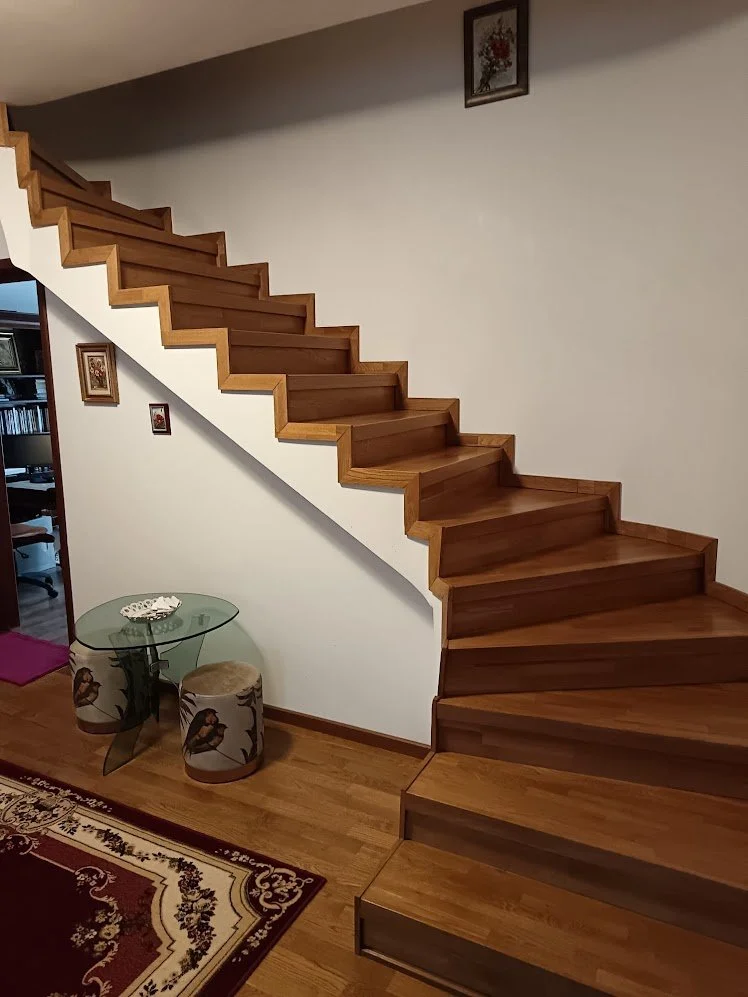 Interior of a home showing a wooden staircase going upstairs along the wall, with a small glass table and two cushioned stools with bird designs nearby. A framed picture hangs above the staircase, and part of a patterned rug is visible on the wooden 