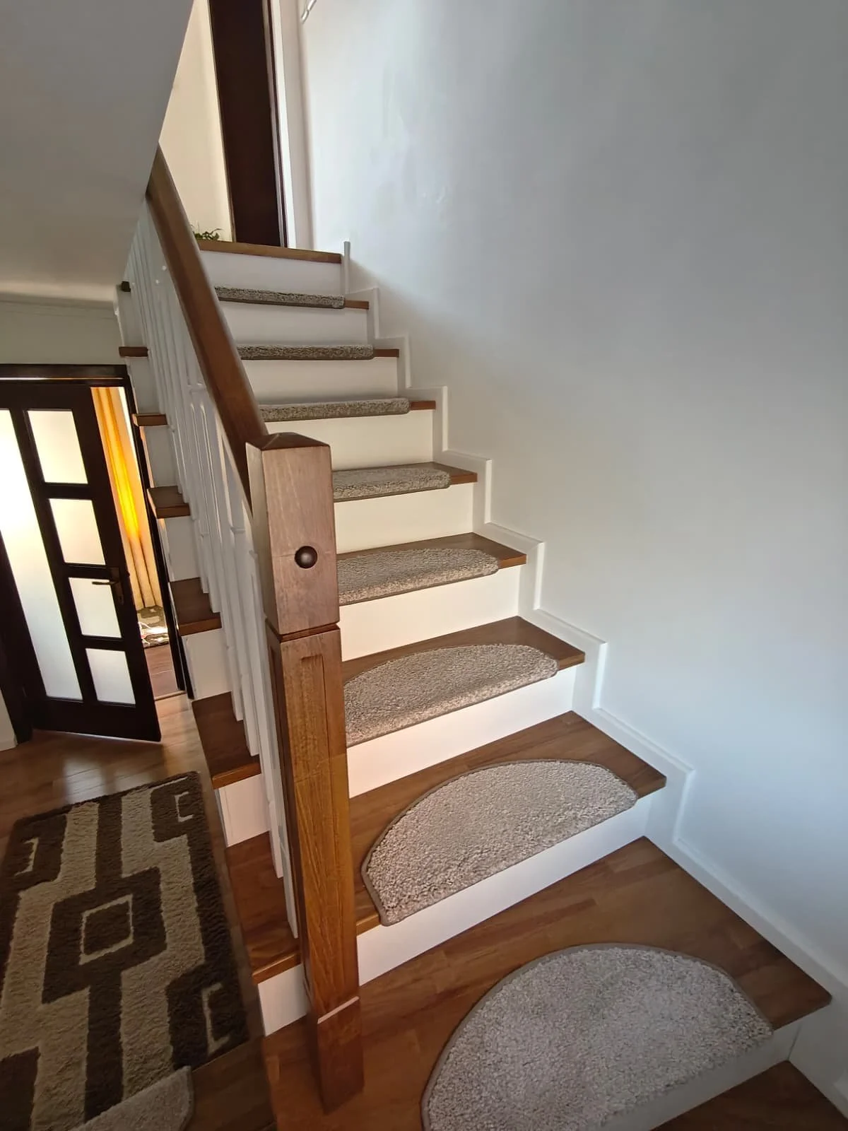 Indoor view of a staircase with beige carpeted steps, wooden railings, and a white wall, leading to a dark door at the top.