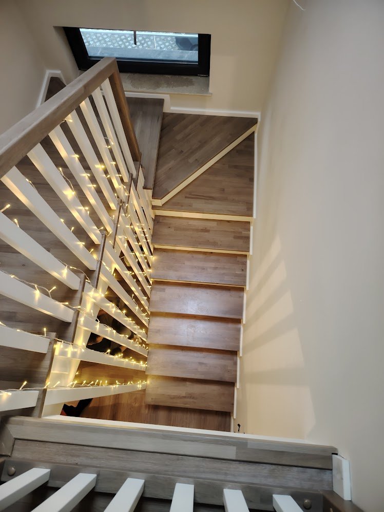 View from the top of a staircase looking down, showing wooden steps with one side decorated with white string lights. There is a small window at the bottom of the stairs.