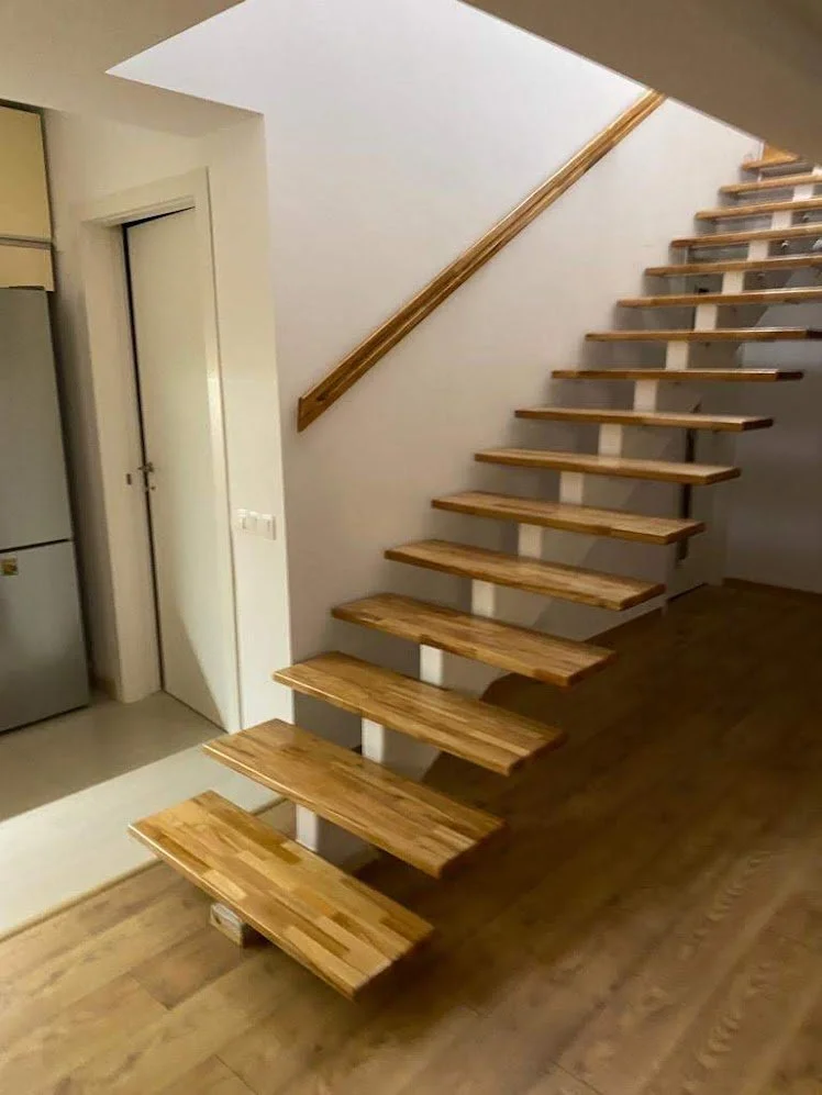 Empty wooden staircase with no supports, leading up to an upper floor, next to a white wall and a wooden handrail.