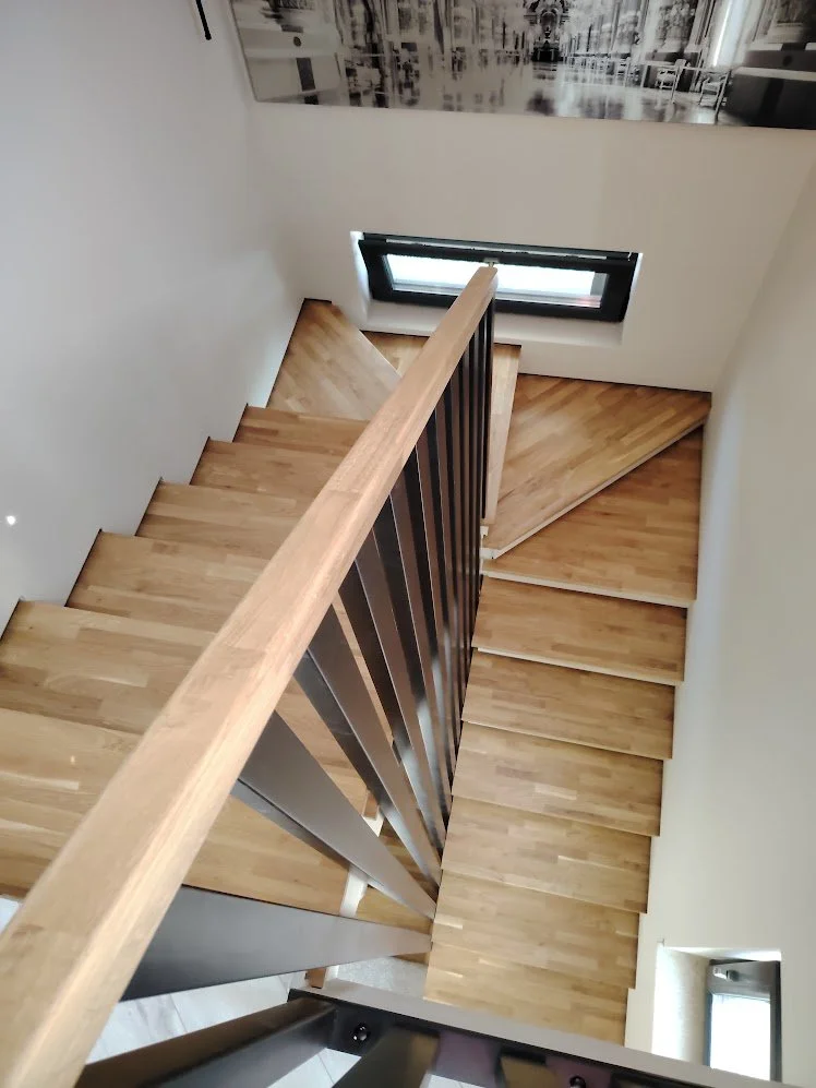 Top-down view of a wooden staircase with a black railing, leading to a lower floor with wooden flooring, near a small window and an abstract black-and-white wall art.