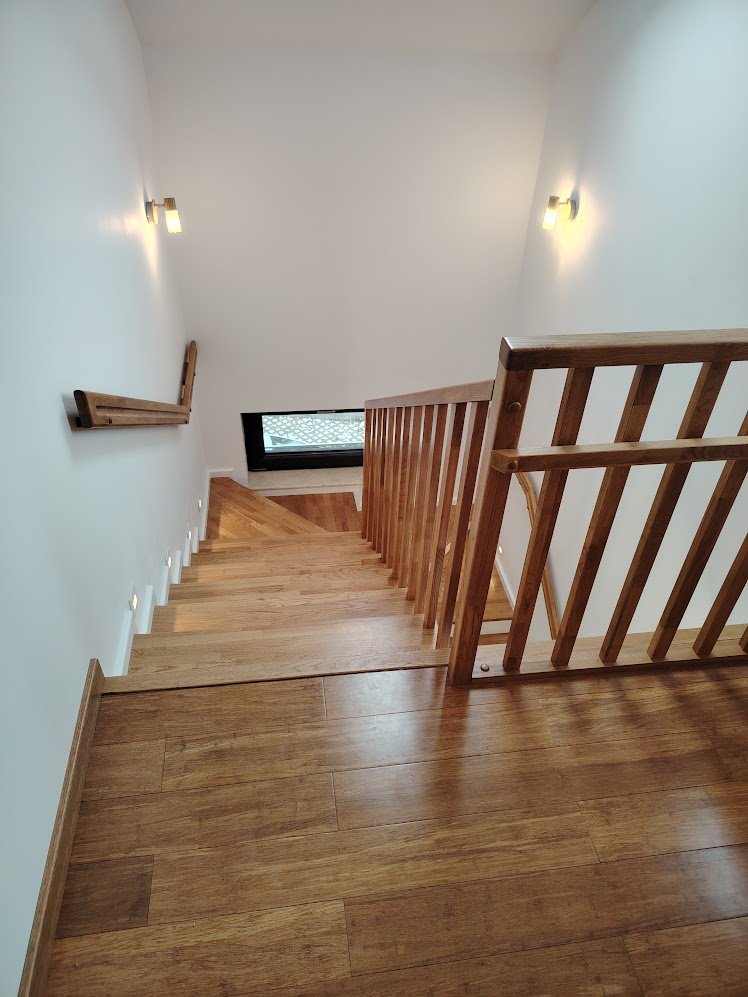 Wooden staircase with railings, viewed from the top, leading down to a lower floor with a window at the bottom of the stairs.