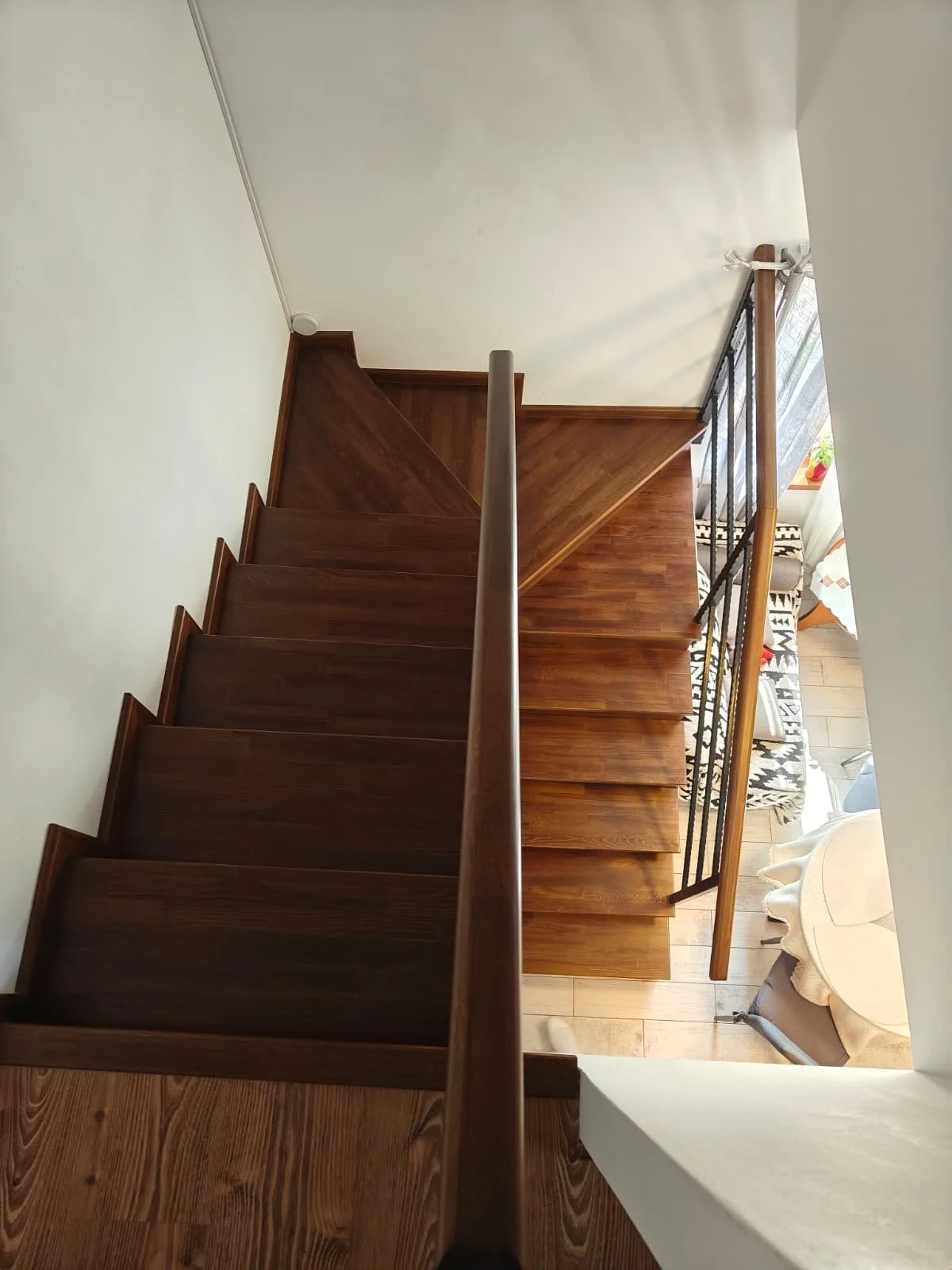 View from top of wooden staircase looking down, with wrought iron and wood railing, living room area with chairs and curtains visible at the bottom.