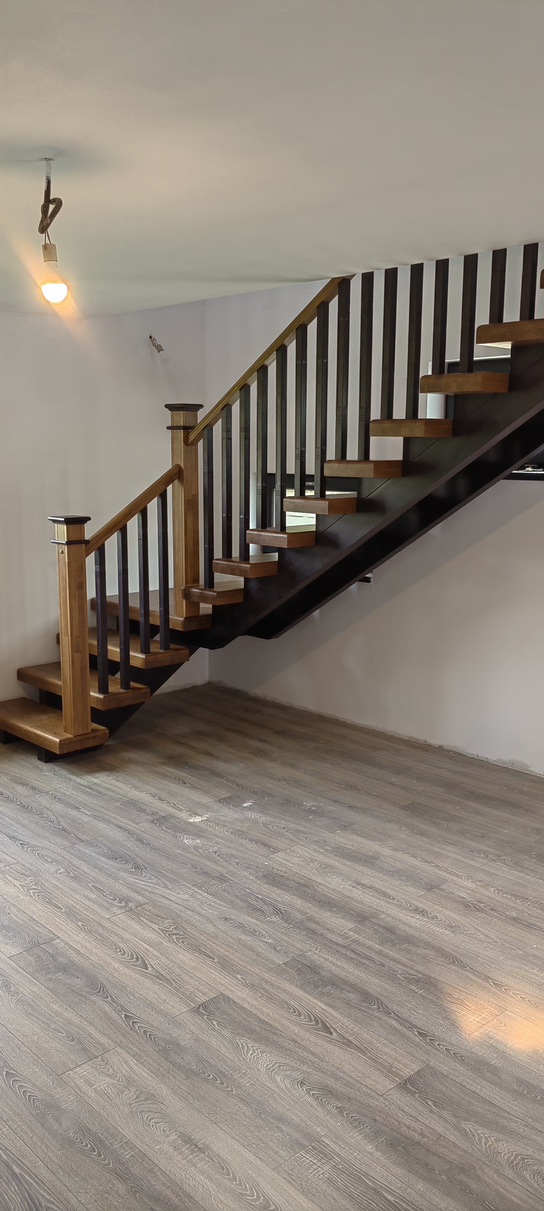 Interior view of a wooden staircase with a black riser, wooden handrails, and black vertical balusters, leading to an upper floor, with a wood-like laminate flooring.