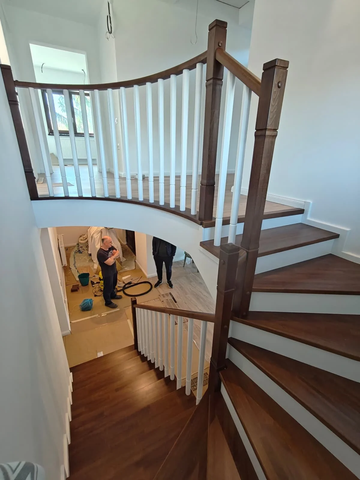 Interior of a house staircase with wooden steps and a white and wood railing, viewed from the top looking down, with people working on home renovation below.