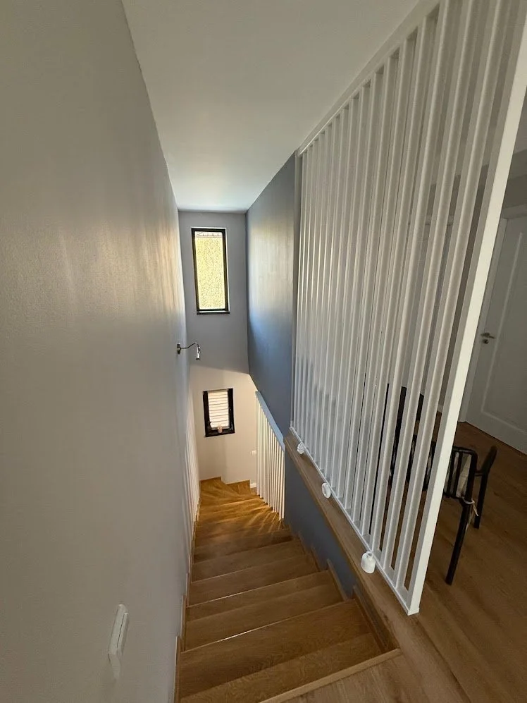 View of a wooden staircase with white railings and a gray wall, leading down to a small landing with two windows, one tall and narrow and the other smaller, allowing natural light into the stairwell.