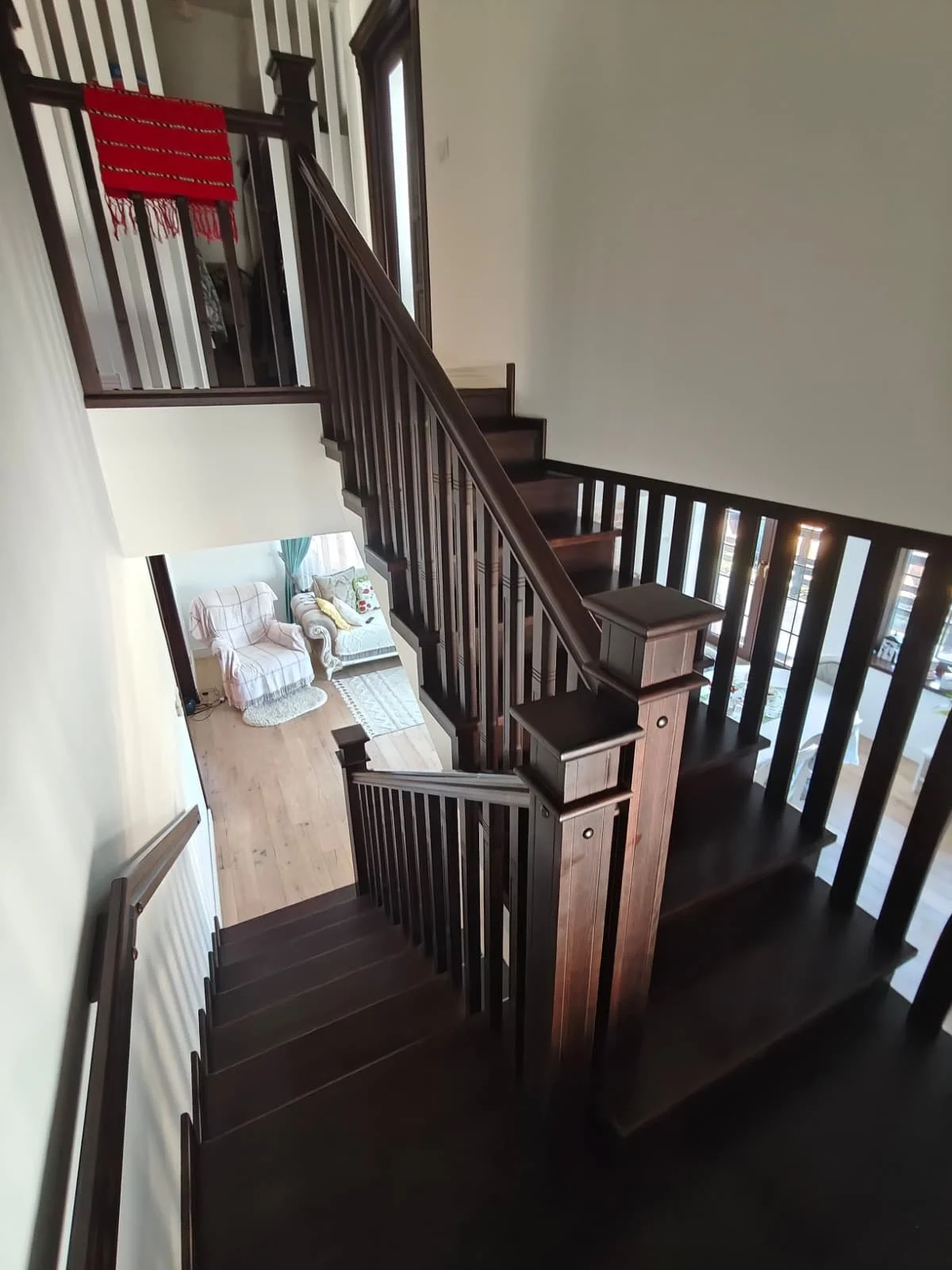 Interior view of a two-story home showing a wooden staircase with dark brown banisters and steps, leading from the second floor down to the living room with white sofas and a small area rug.