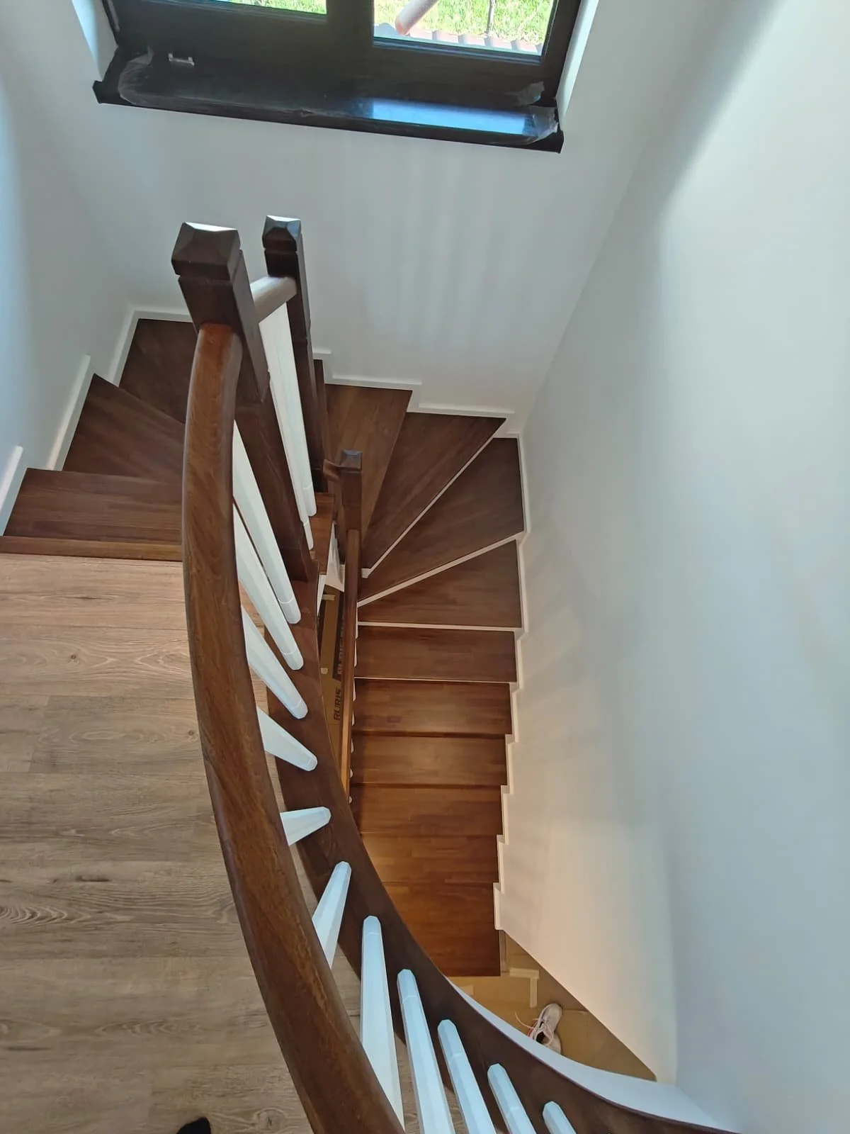 Top view of a wooden staircase with a handrail, white balusters, and a window at the top letting in natural light.