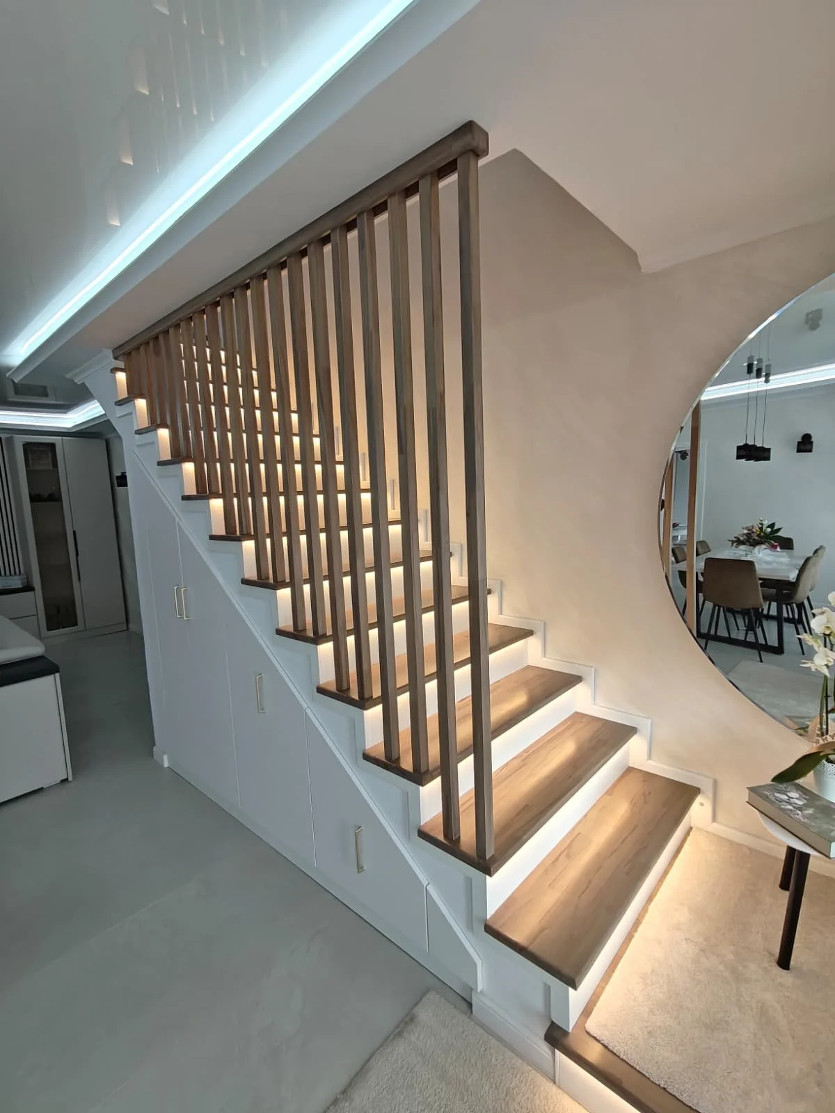 Interior view of a modern staircase with wooden steps and vertical wooden balusters, integrated with under-stair storage cabinets, and a circular mirror reflecting a dining area with a table and chairs.