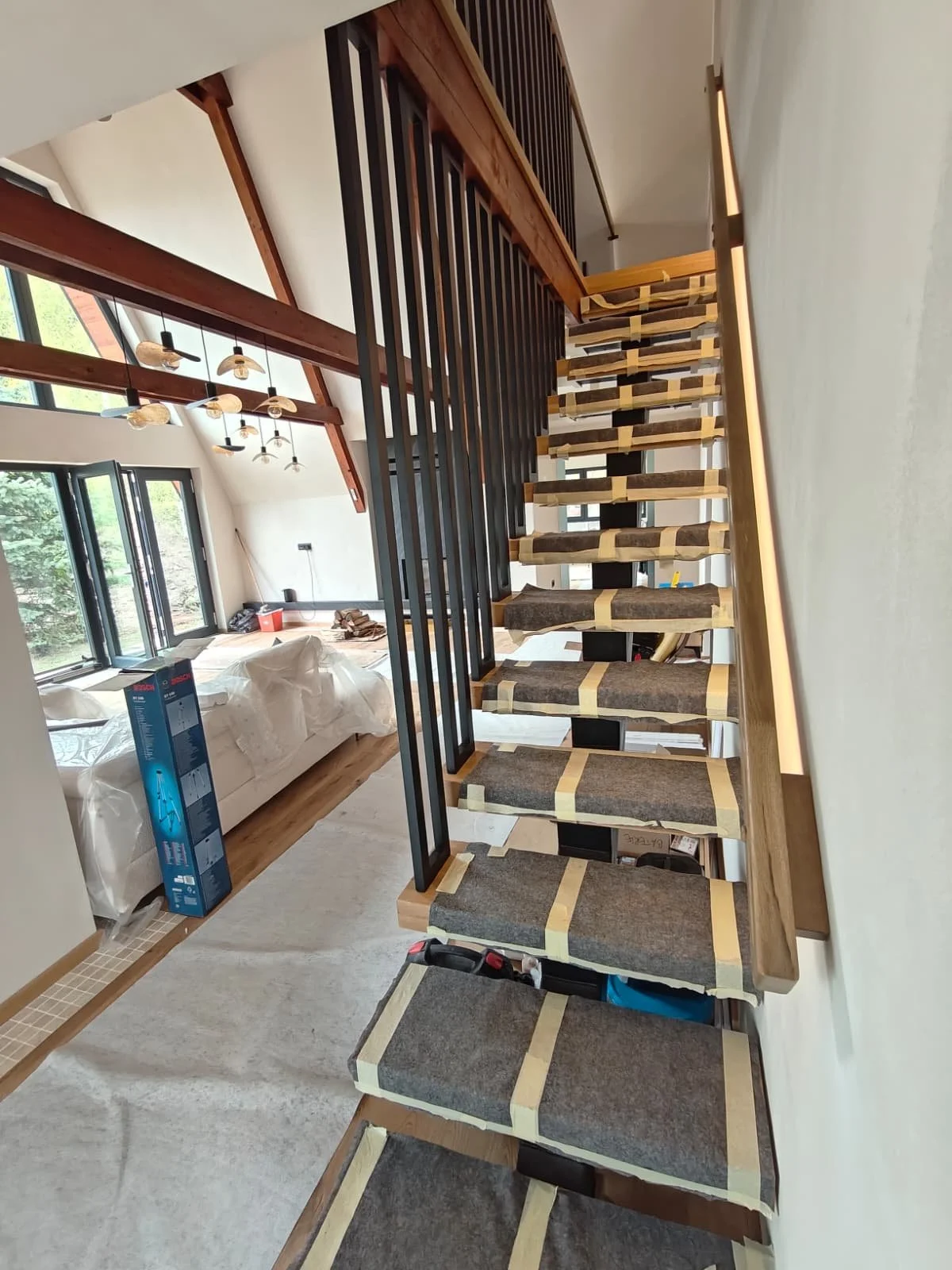 The interior of a house under construction, showing a staircase with cardboard and tape on the steps, a living room with large windows, a couch covered with plastic, and various construction materials and tools.