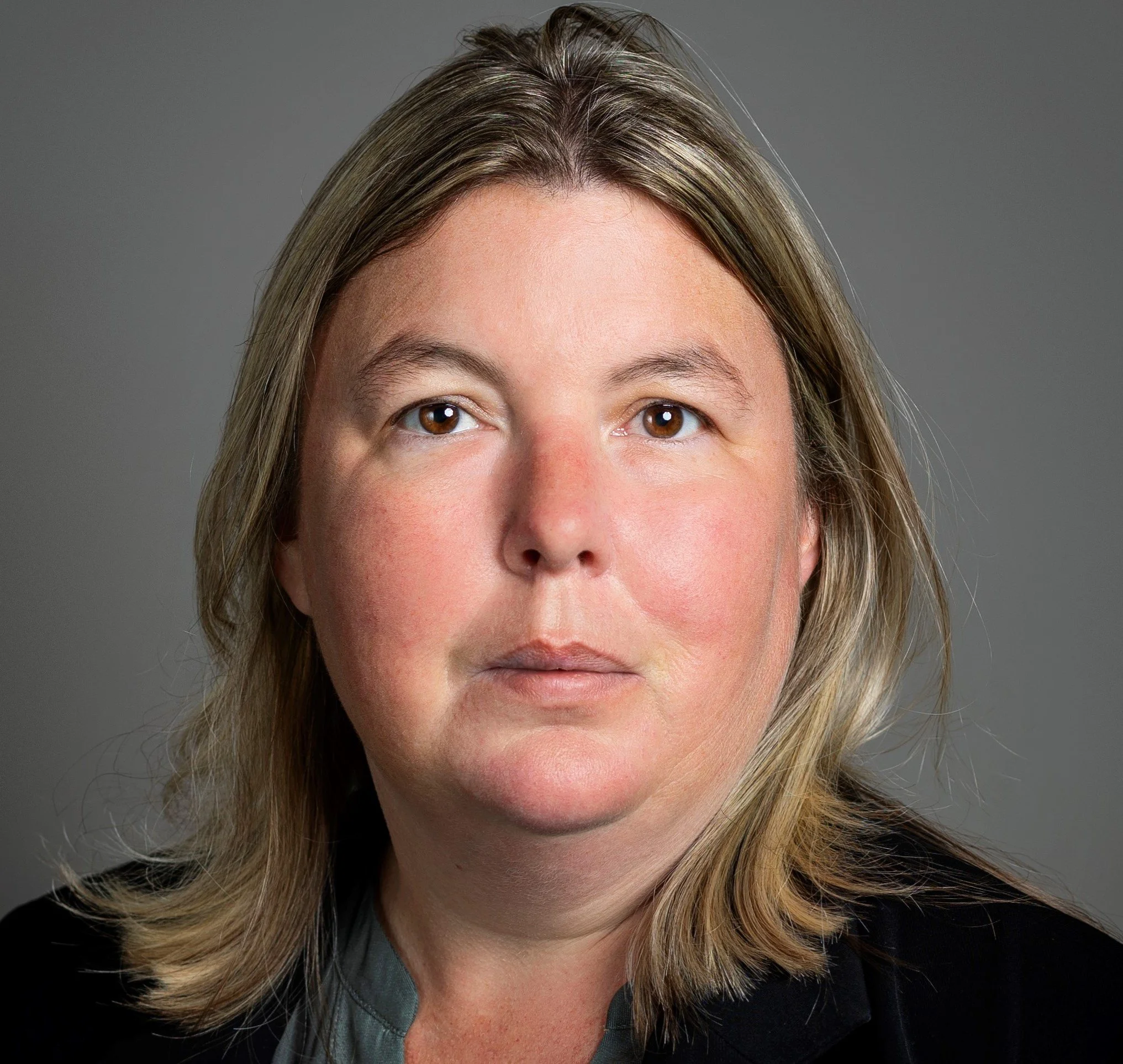 Close-up portrait of a woman with shoulder-length blonde hair, brown eyes, wearing a black blazer and gray shirt, against a plain gray background.