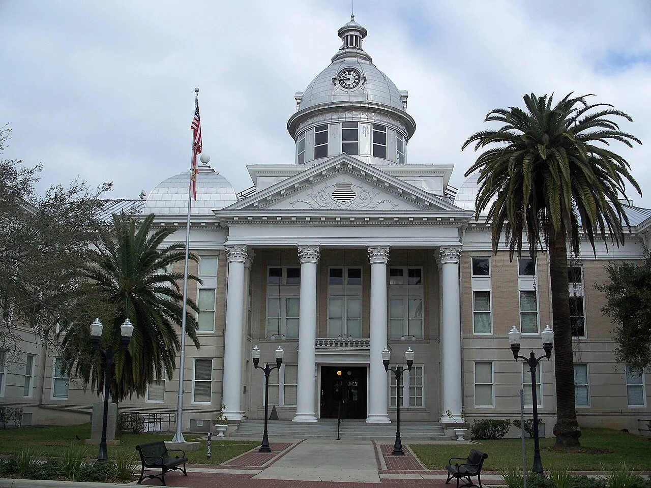 A historic white courthouse with tall columns, a dome with a clock, and surrounded by palm trees, benches, and lamp posts.