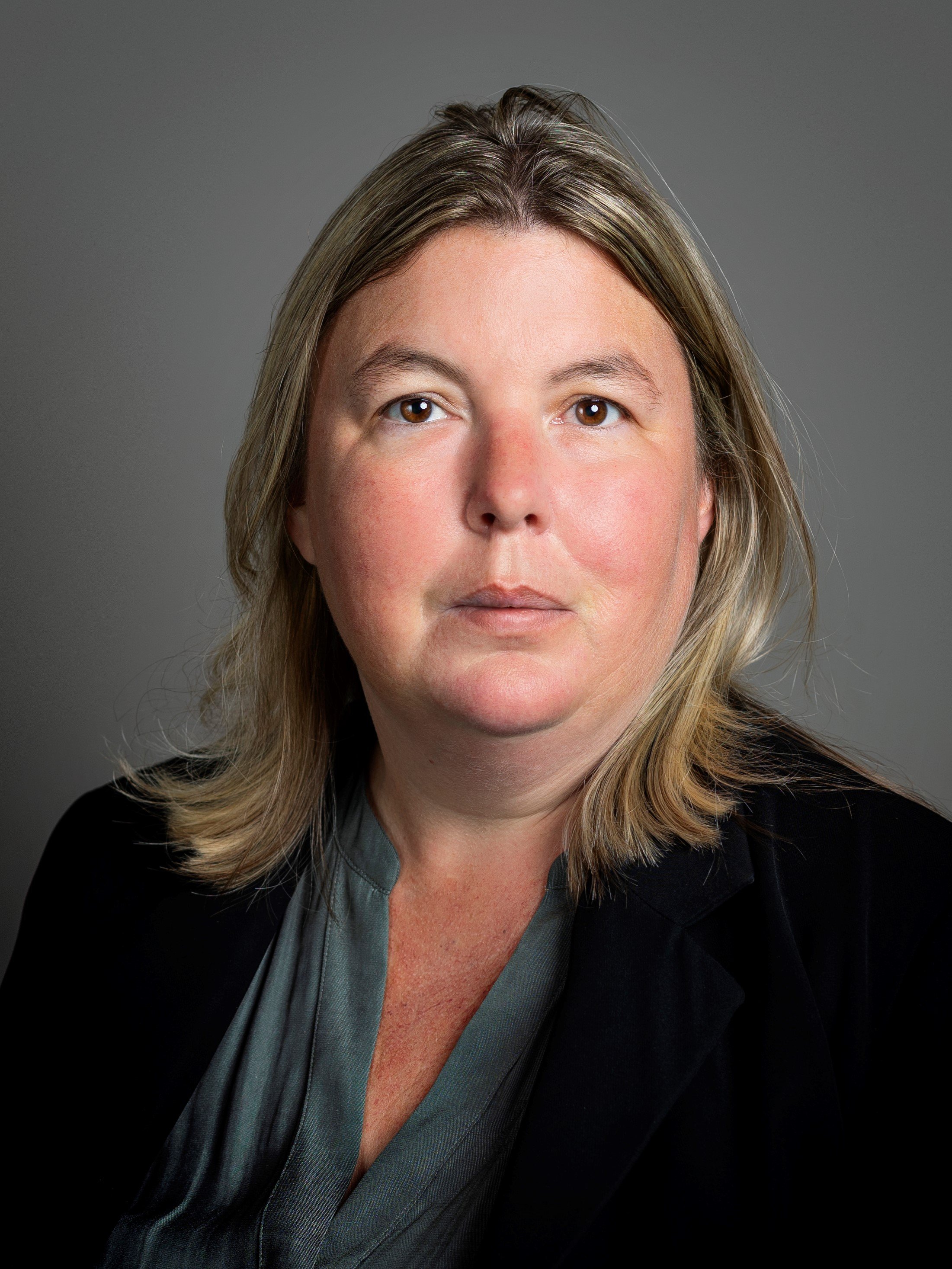 A woman with shoulder-length blonde hair wearing a dark blazer over a gray blouse, looking directly at the camera with a neutral expression.