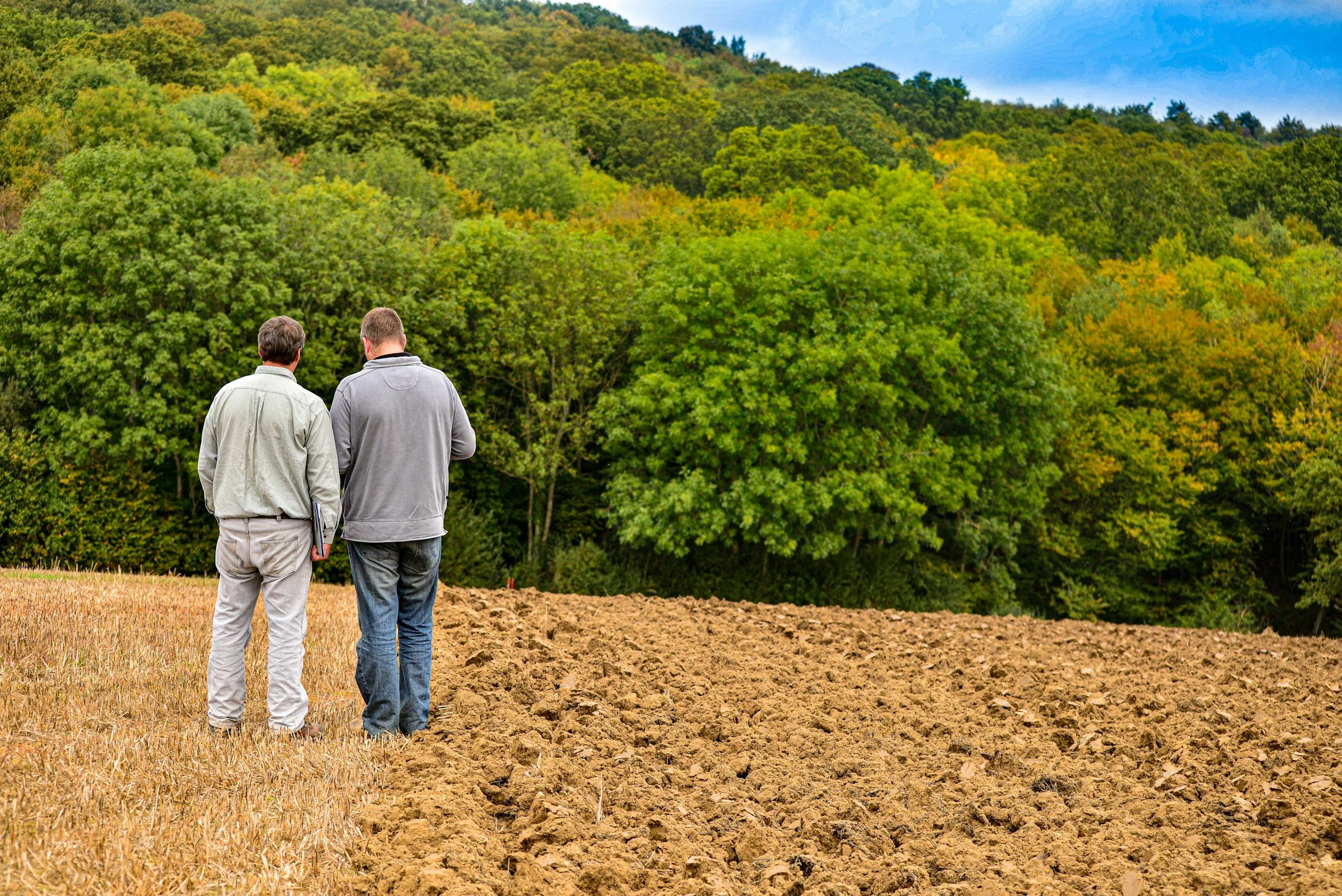Two men walking through a field of tilled soil towards a green forested hillside under a partly cloudy sky.