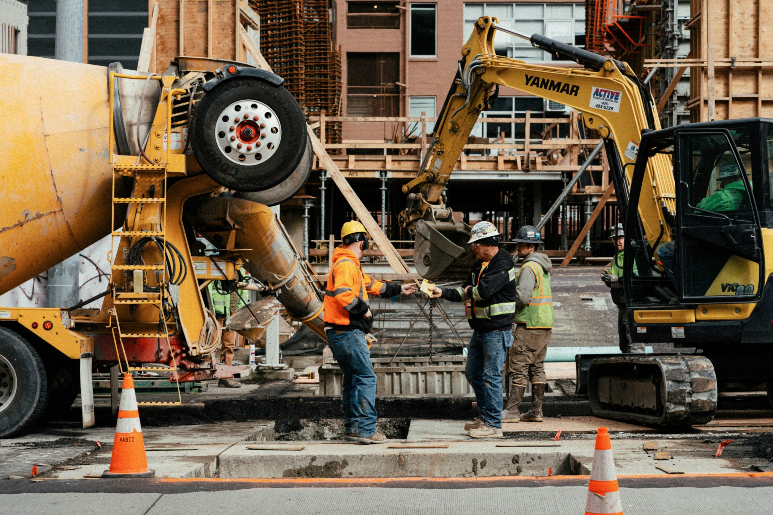 Construction workers wearing helmets and safety vests work on a building site with heavy machinery, including a cement mixer and excavator, amid orange traffic cones and wooden scaffolding.