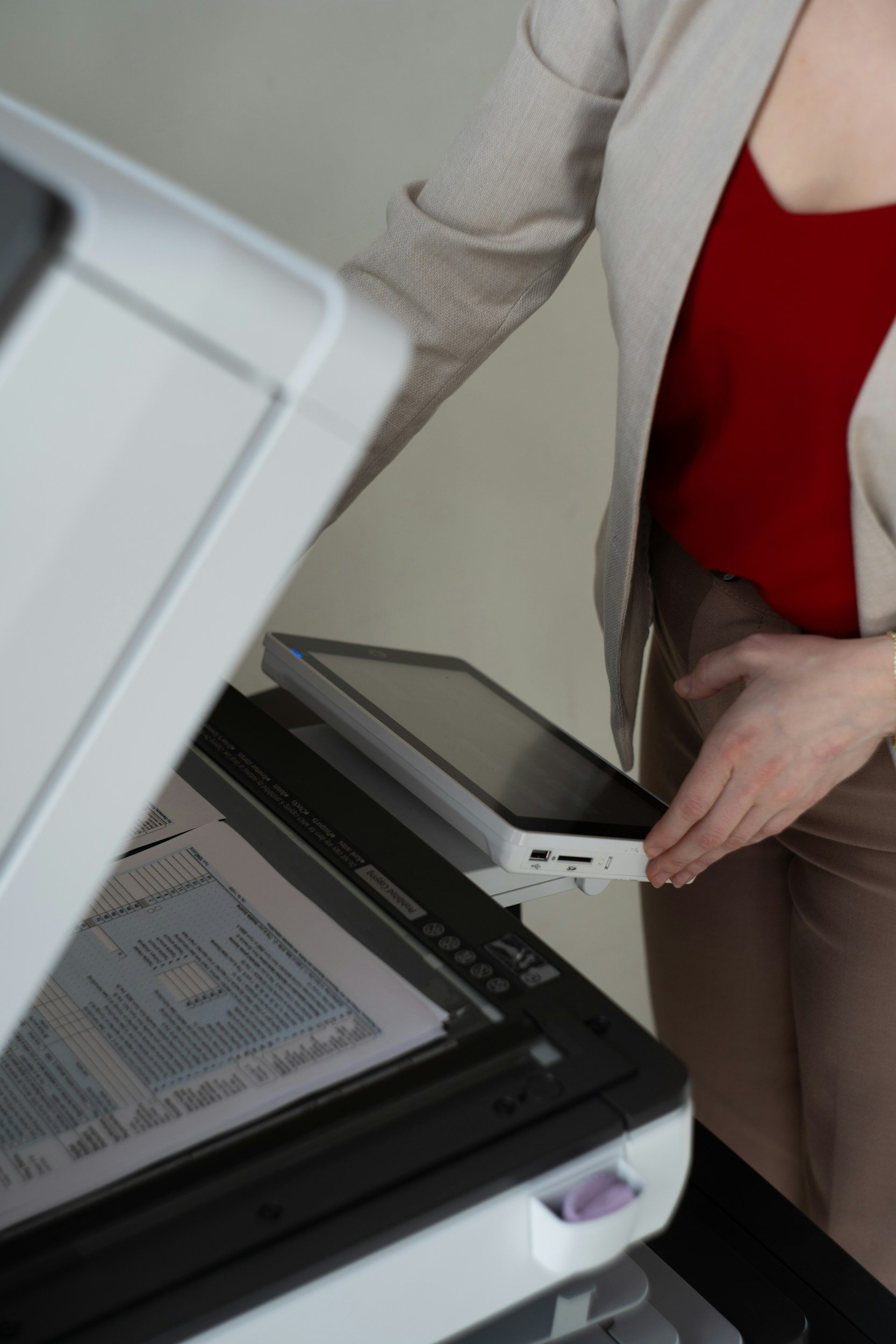 A person wearing a beige blazer and red top is using a barcode scanner on a sheet of paper at a desk with various documents and office supplies.