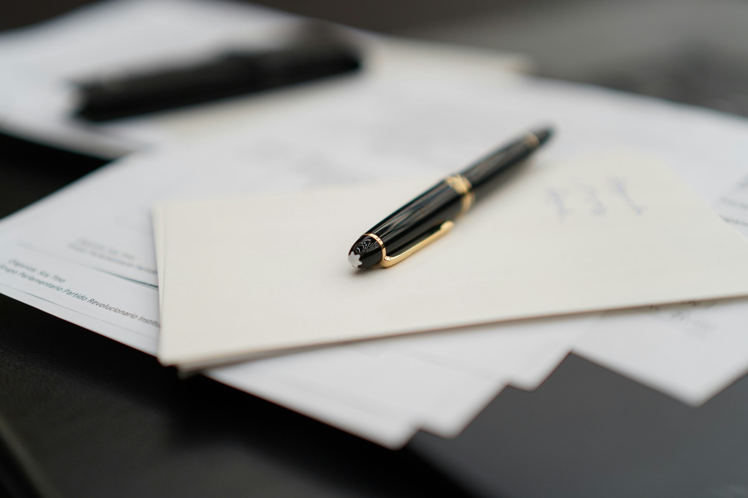 A close-up of a black and gold pen resting on a white sheet of paper, with blurred documents and objects in the background.