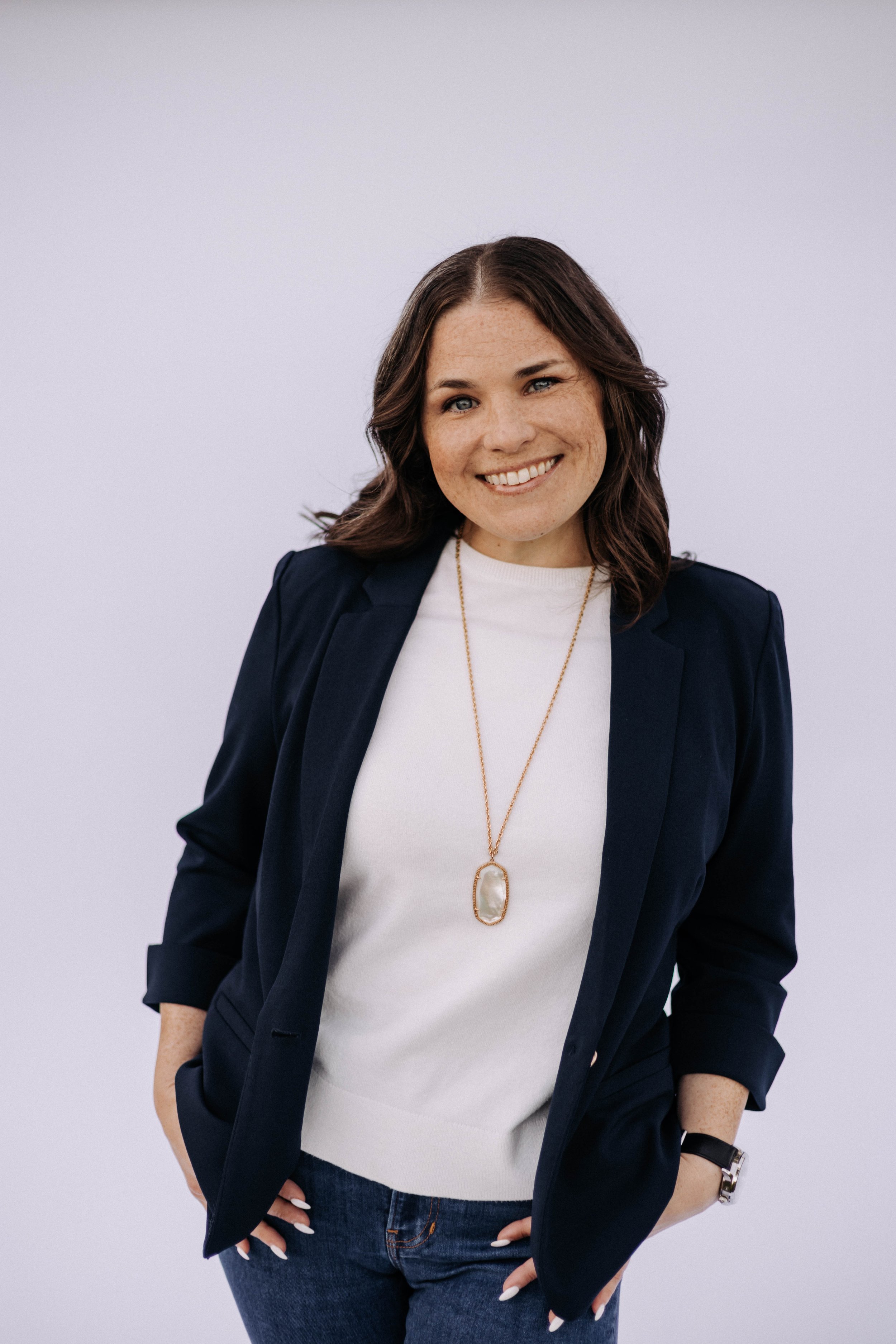 A woman with medium-length wavy brown hair, blue eyes, and freckles, smiling and wearing a navy blazer over a white shirt, paired with dark jeans, accessorized with a gold necklace with a large oval pendant, a black watch, and a silver bracelet.