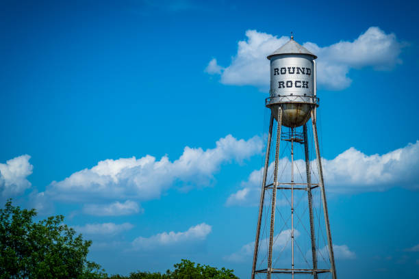 A tall, metal water tower with a round top, labeled 'ROUND ROCK', stands against a bright blue sky with scattered clouds. Green trees are visible at the bottom of the image.