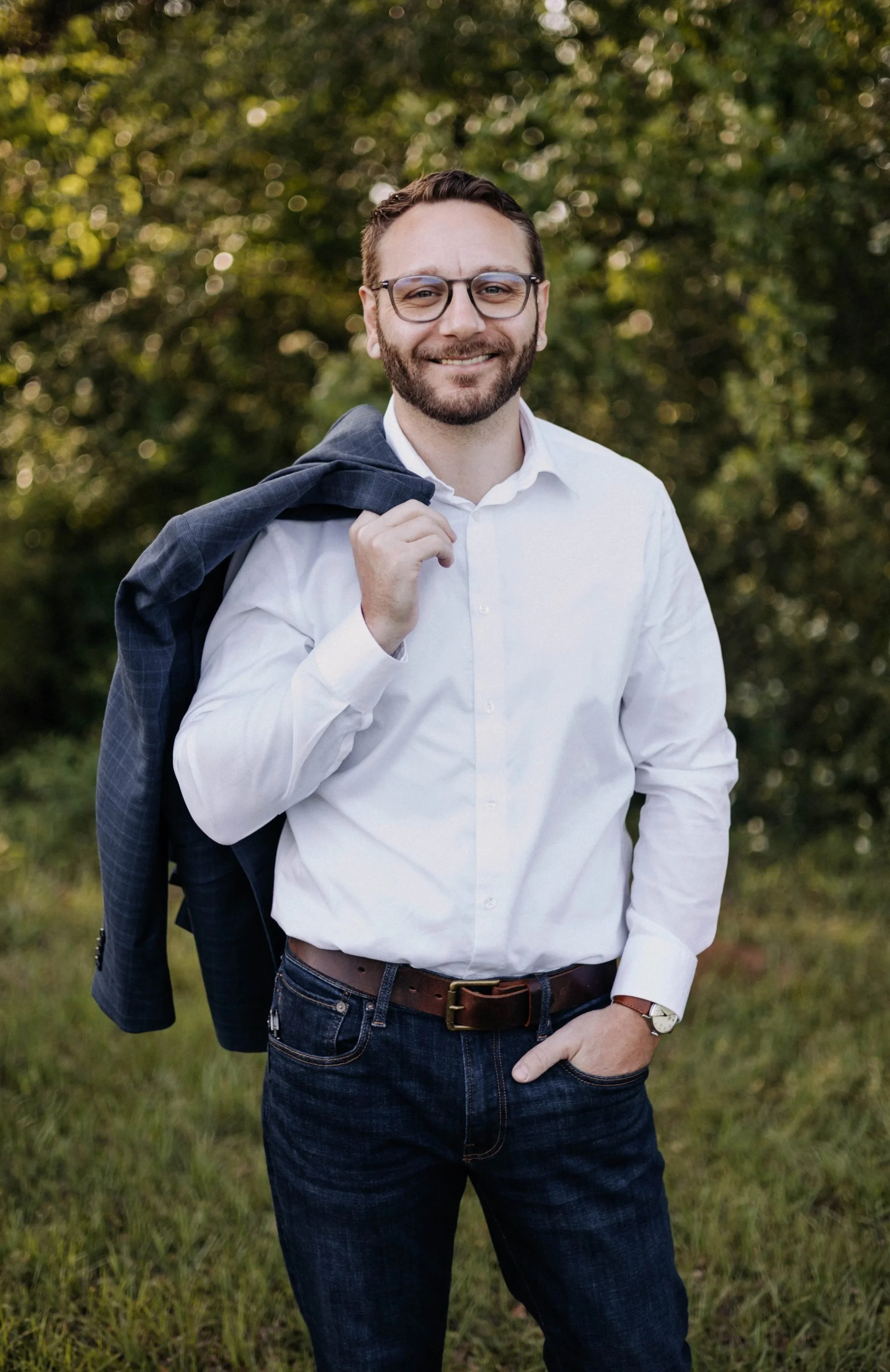 A smiling man with glasses, wearing a white button-up shirt, blue jeans, and a wristwatch, holding a blazer over his shoulder in a park with green trees in the background.