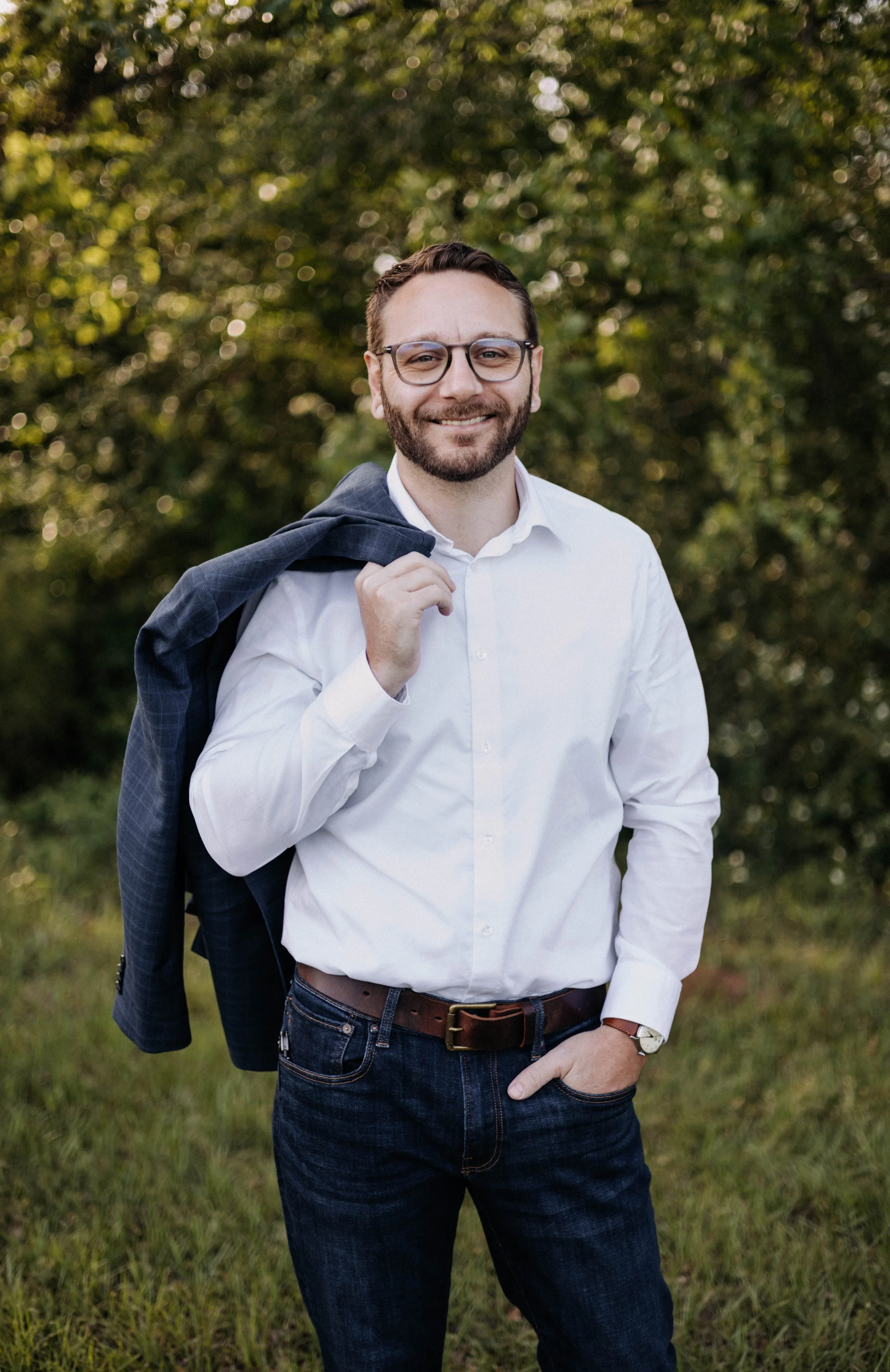 A man with glasses and a beard smiling confidently outdoors, holding a dark blazer over his shoulder, wearing a white shirt and dark jeans, with a green, leafy background.