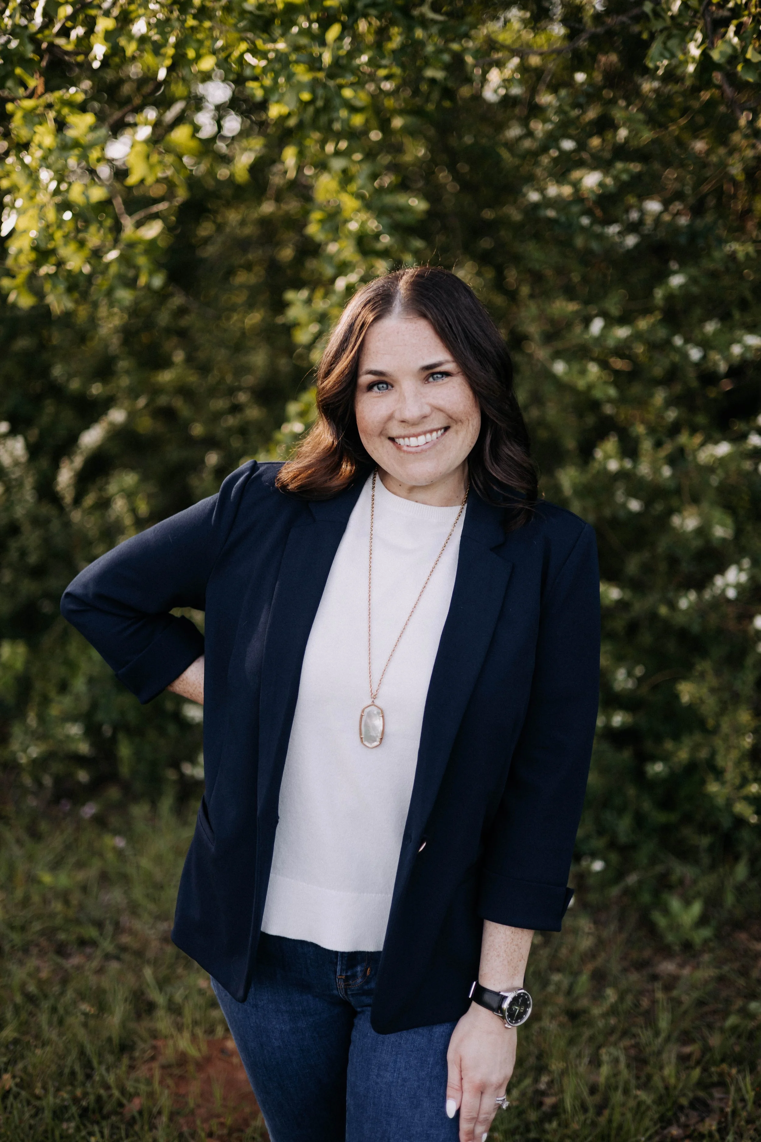 A woman with shoulder-length dark hair and light skin standing outdoors in front of greenery, smiling with her hand on her hip. She is wearing a dark blazer, white shirt, blue jeans, a watch, and a necklace with a large pendant.