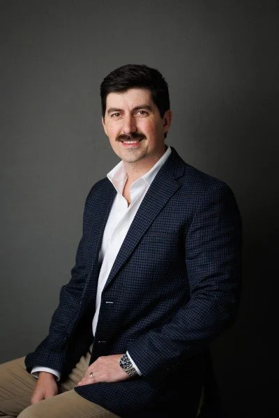 A man with dark hair and a mustache wearing a navy checked blazer, white shirt, and beige pants, posing against a gray background.