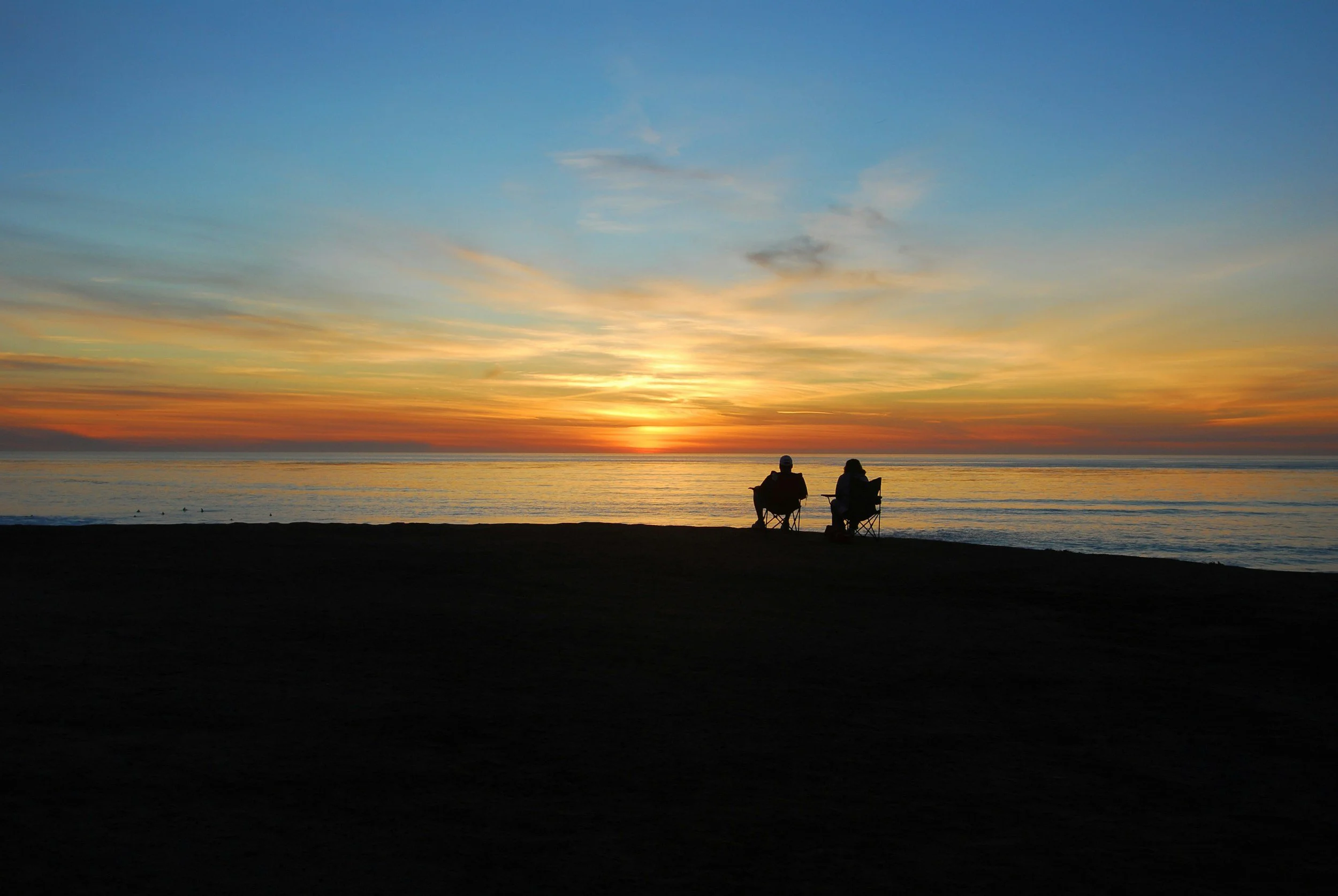 Two people sitting on chairs on a beach during sunset, with colorful sky and calm ocean.