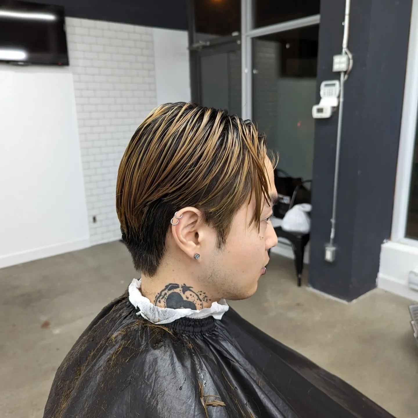 A person with short, styled hair with blonde highlights and dark roots, sitting in a hair salon. The person has multiple earrings, including a helix piercing and a stud. They are wearing a black cape with a white paper collar at the neck, and a tatto