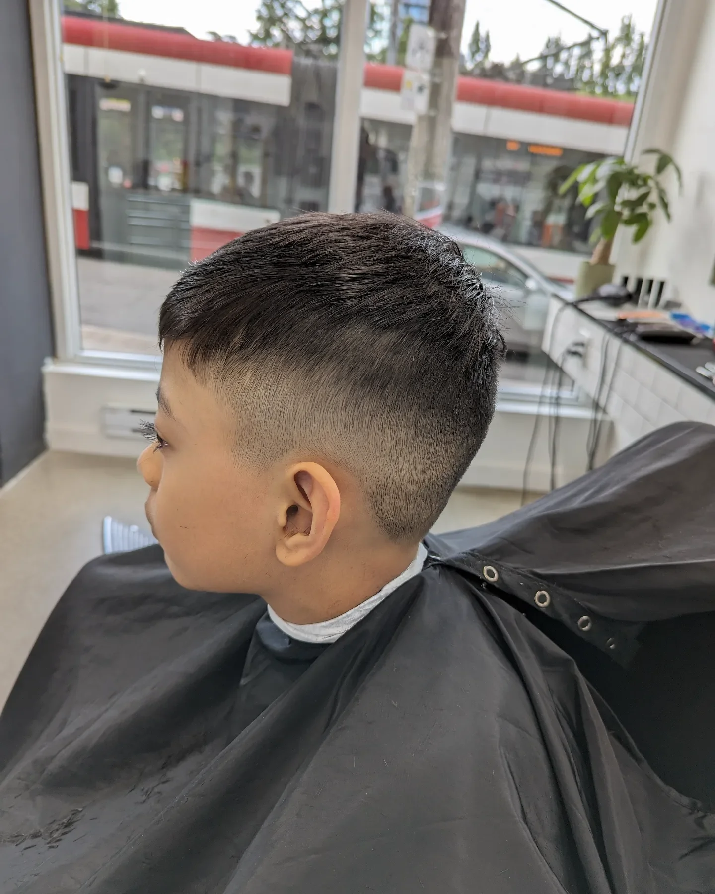 Side view of a young boy with a fresh fade haircut sitting in a salon chair, with large windows showing a city bus outside.