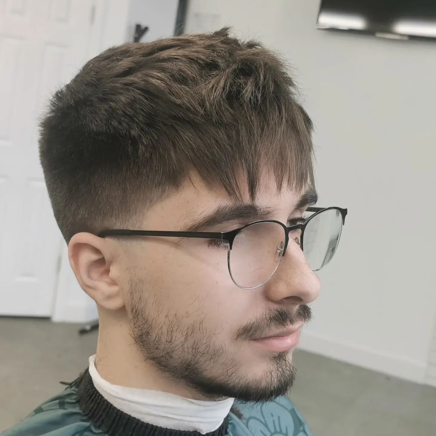 A young man with glasses, a short beard, and a fresh haircut, sitting in a barbershop.