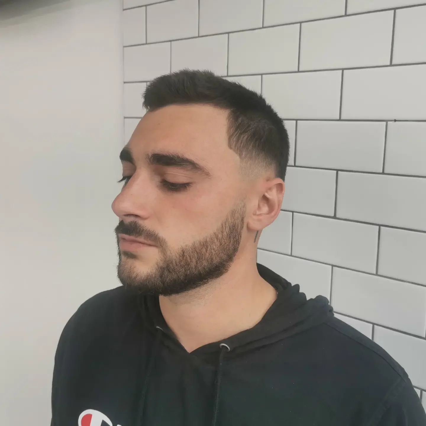 A man with a short, dark, styled haircut and a beard, wearing a black hoodie, standing against a tiled wall with his eyes closed.