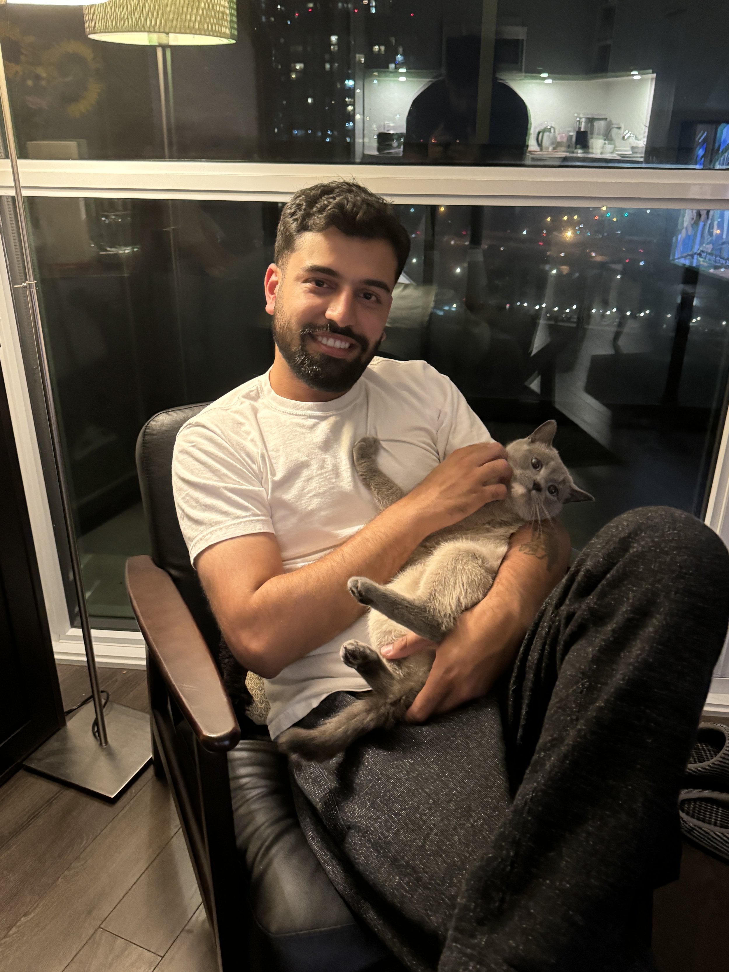 A man with dark hair, a beard, and light skin wearing a white T-shirt, smiling while sitting in a black armchair at night. He is holding a gray cat on his lap, petting it with one hand. Behind him, a large window reveals city lights and a distant building. To his left, there is a tall green lamp and a small bar area with various bottles and kitchen appliances.