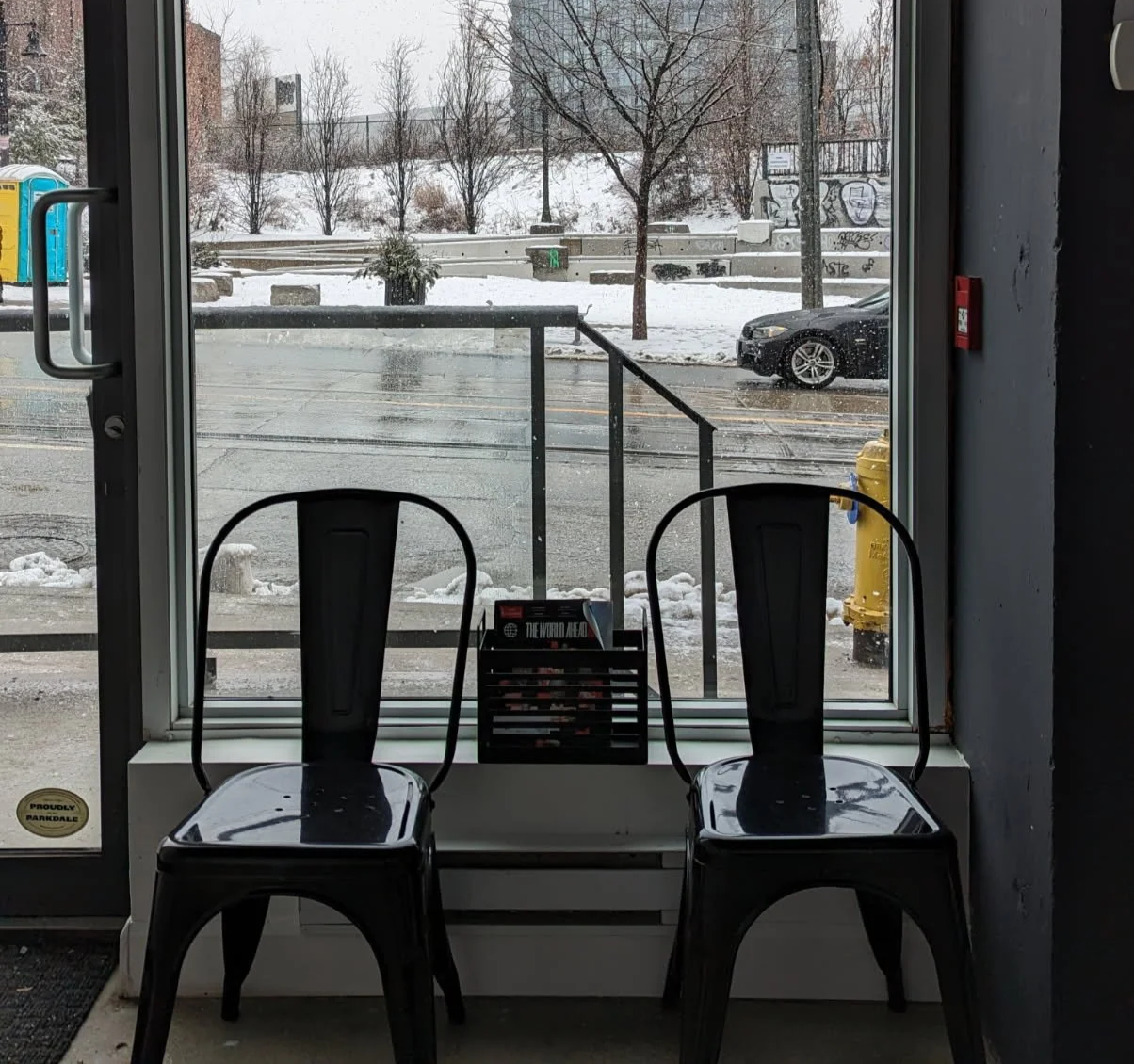 View through a glass door showing two black chairs inside, a window with a snowy street scene outside including a black car, trees, graffiti on a wall, and a yellow fire hydrant.