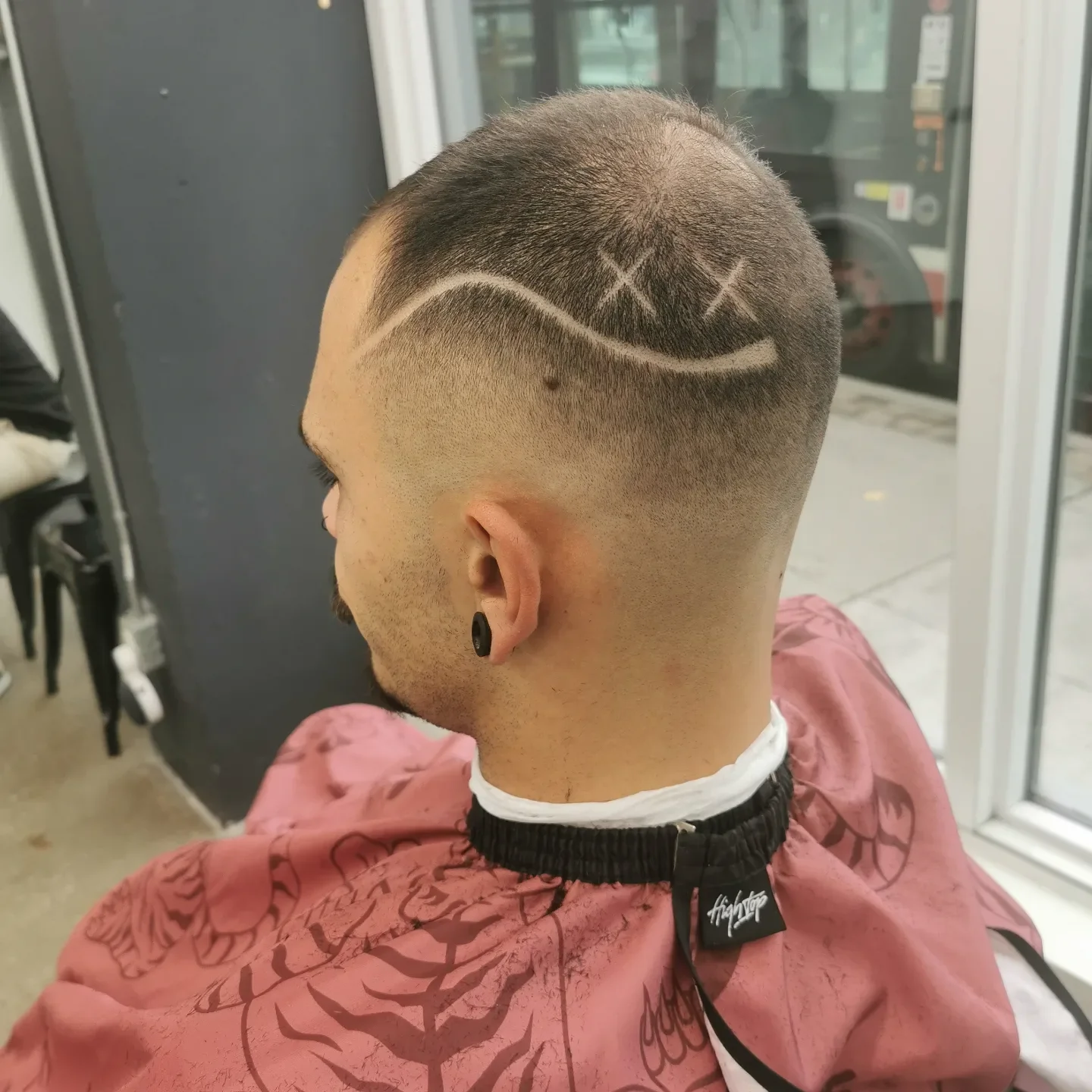 A man with a textured haircut featuring a wave design and three X's shaved into the top of his head, sitting in a barber's chair wearing a pink barber cape.