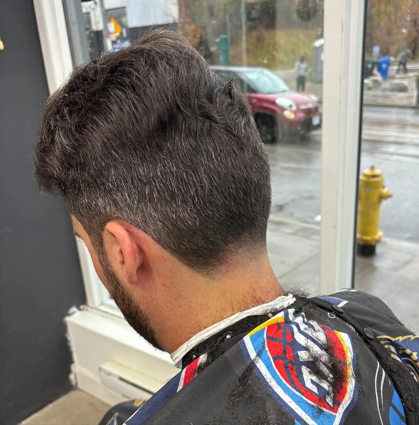 Man sitting in barber's chair with freshly cut short hairstyle, facing away from camera. Outside view through window shows street scene with cars and pedestrians on a rainy day.