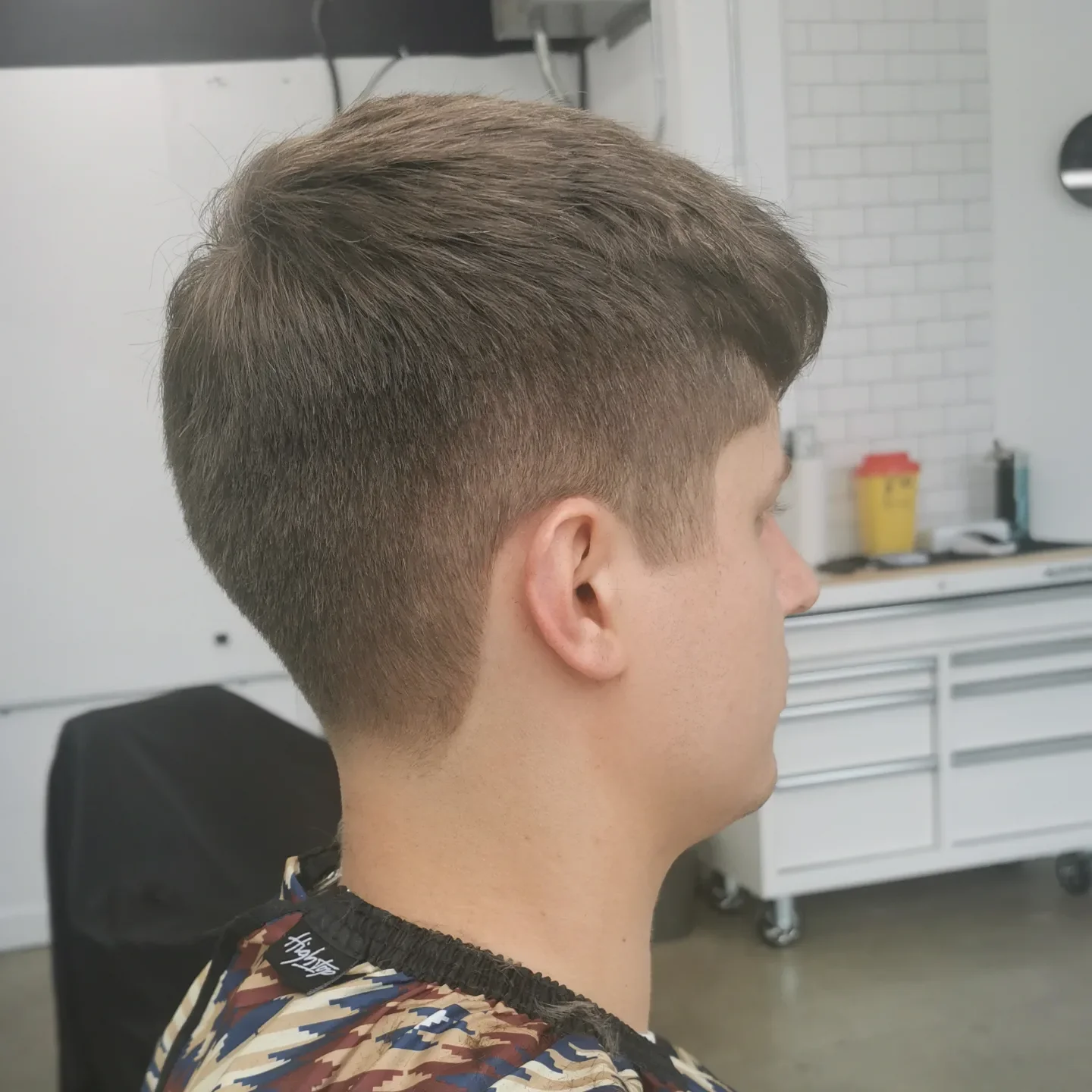 Side view of a young man with a new haircut, featuring short sides and longer top hair, in a barbershop.