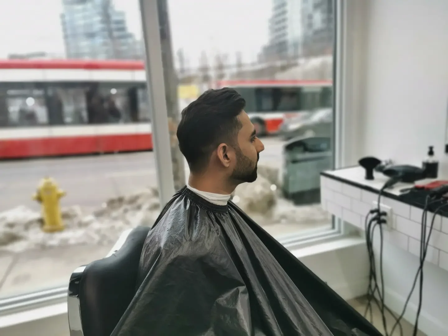 Man with dark hair and beard getting a haircut at a barbershop, sitting in a black cape, with a large window behind him showing city street and bus outside.