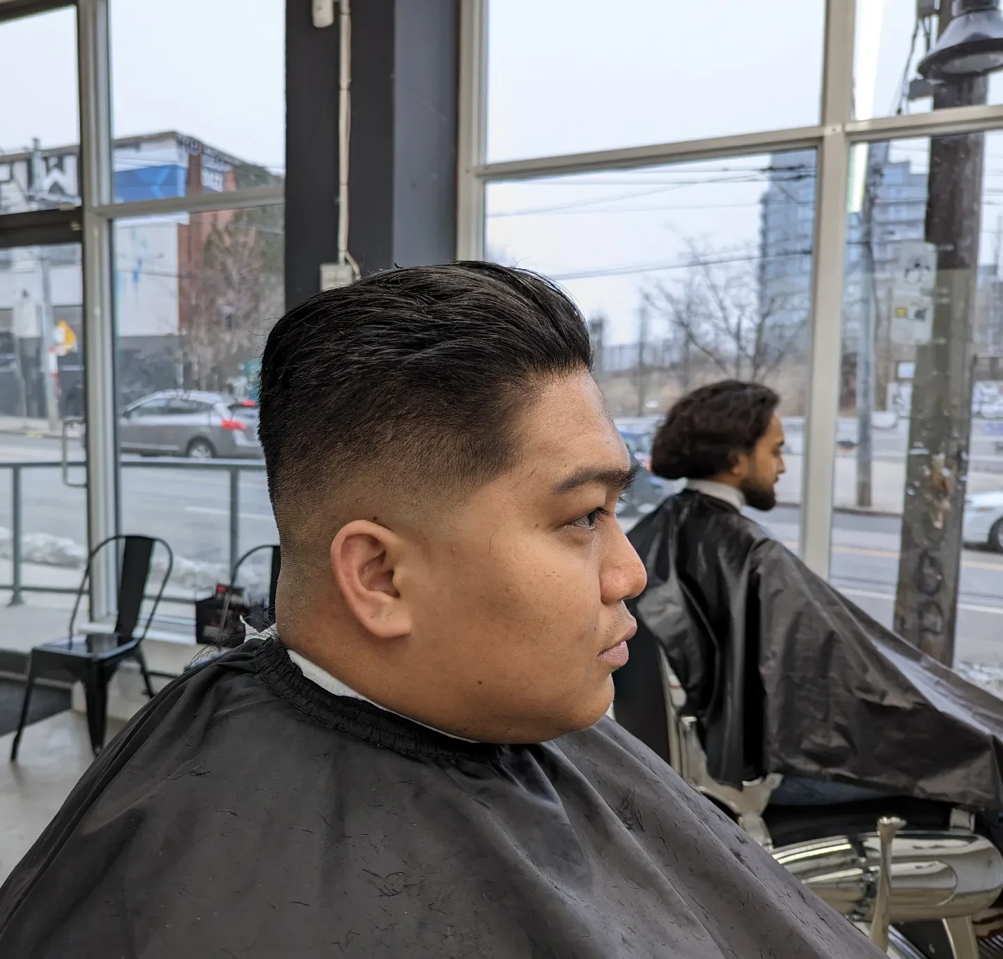 Man with a fresh fade haircut sitting in a barber shop, looking to the side, with another man in the background also seated in a barber chair.