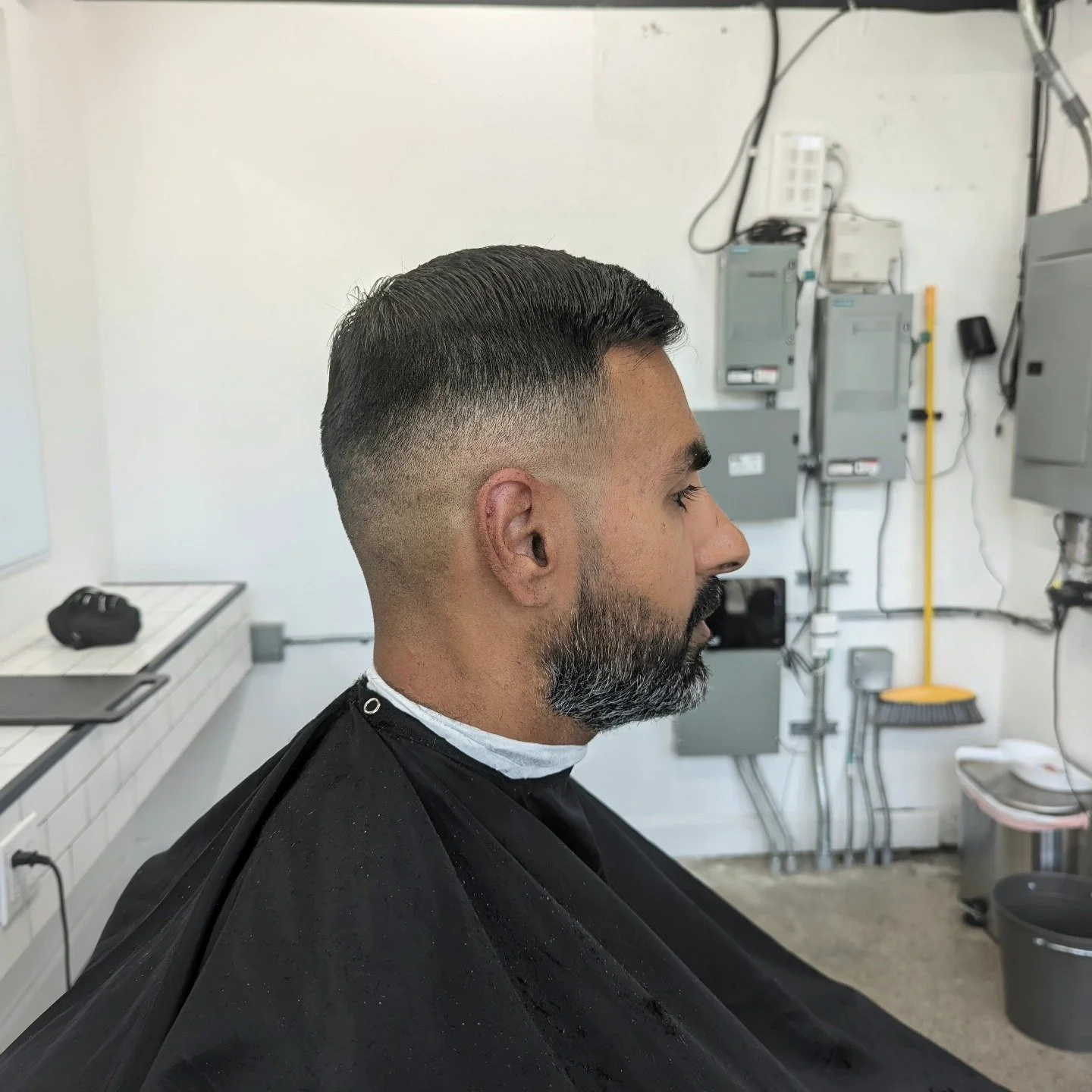 Side view of a man sitting in a barbershop chair, sporting a fresh haircut with a fade and a beard, with a white wall and electrical equipment in the background.