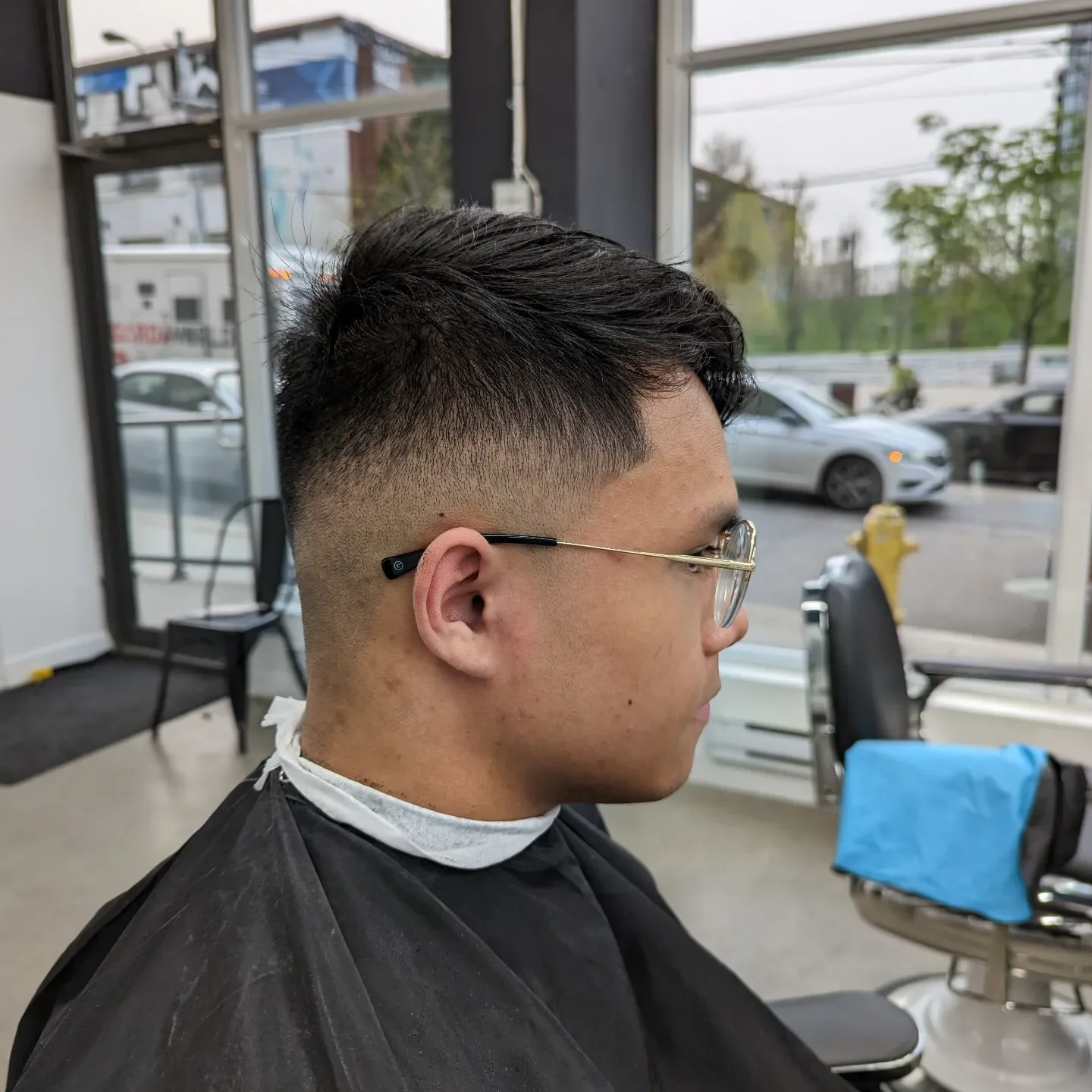 A man with dark hair and glasses getting a fade haircut in a barbershop, sitting in a chair with a black cape, inside a shop with large windows showing parked cars and a street outside.